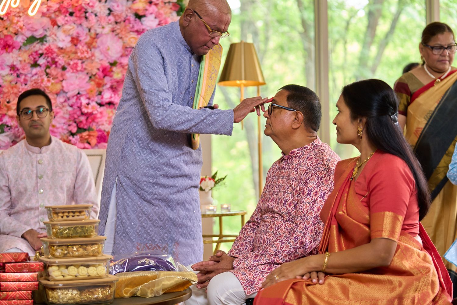 Elderly man in a lavender kurta blesses a seated couple during a traditional Indian ceremony with floral decor and ceremonial trays.