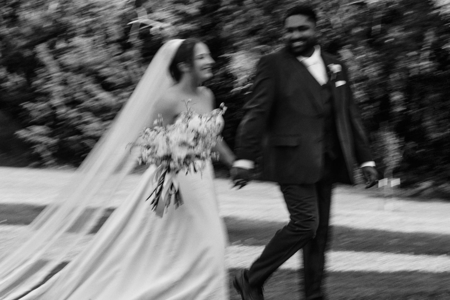 Black and white photo of a smiling bride in a flowing veil and gown holding a bouquet while walking hand in hand with her groom outdoors.