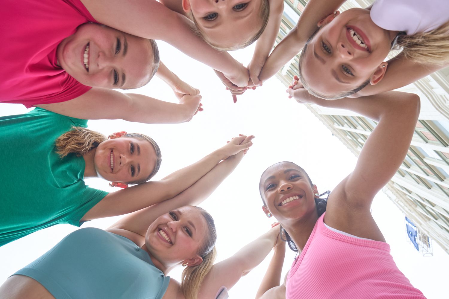 Five smiling women in colorful athletic wear huddle together in a circle, viewed from below against a bright sky.