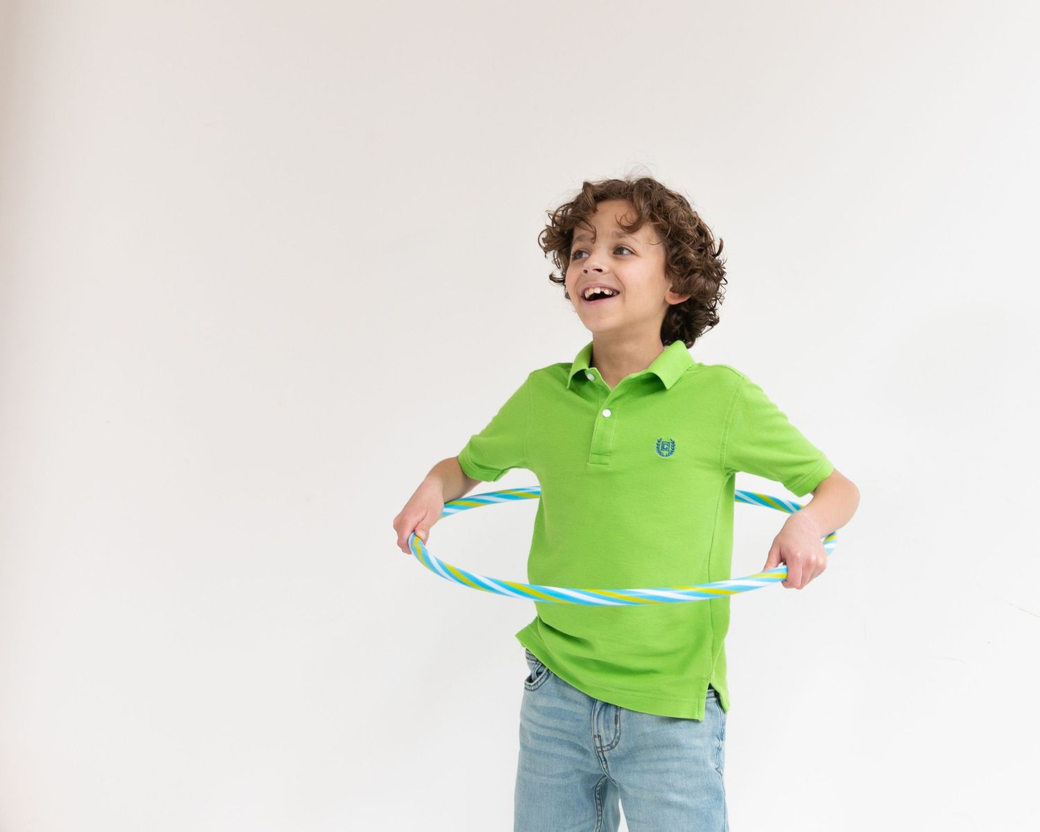 A cheerful boy in a green polo shirt plays with a blue hula hoop against a white background.