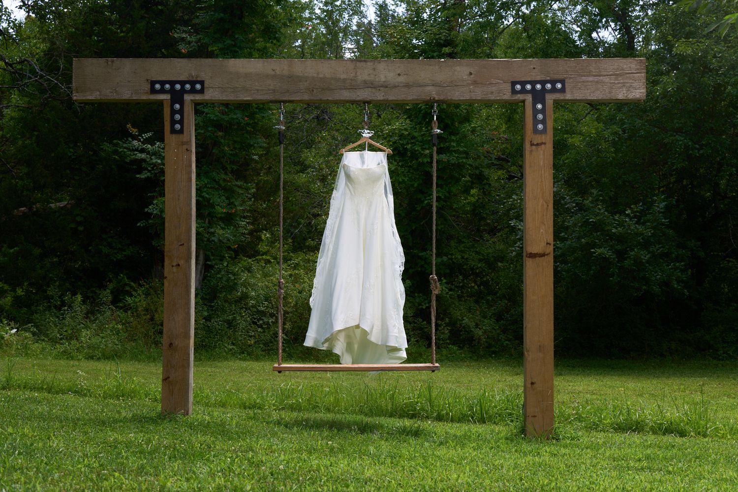White wedding dress hanging from a rustic wooden swing frame on a green lawn with lush trees in the background.