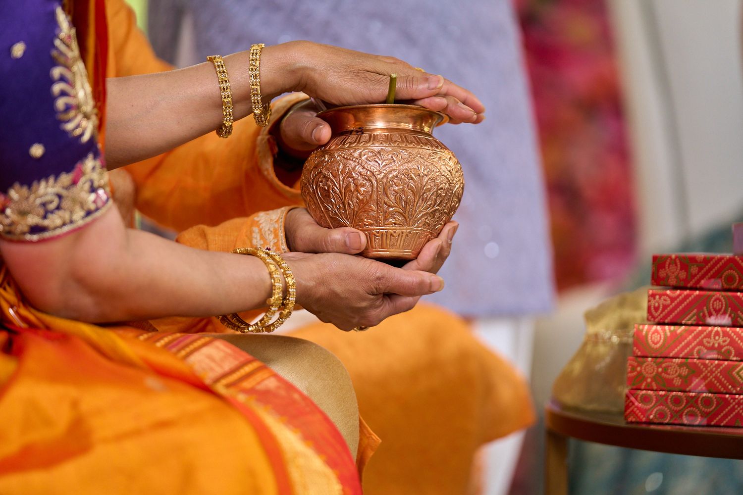 Woman in an orange silk saree and gold bangles holds an ornate copper kalash pot with both hands during a traditional Indian ceremony.