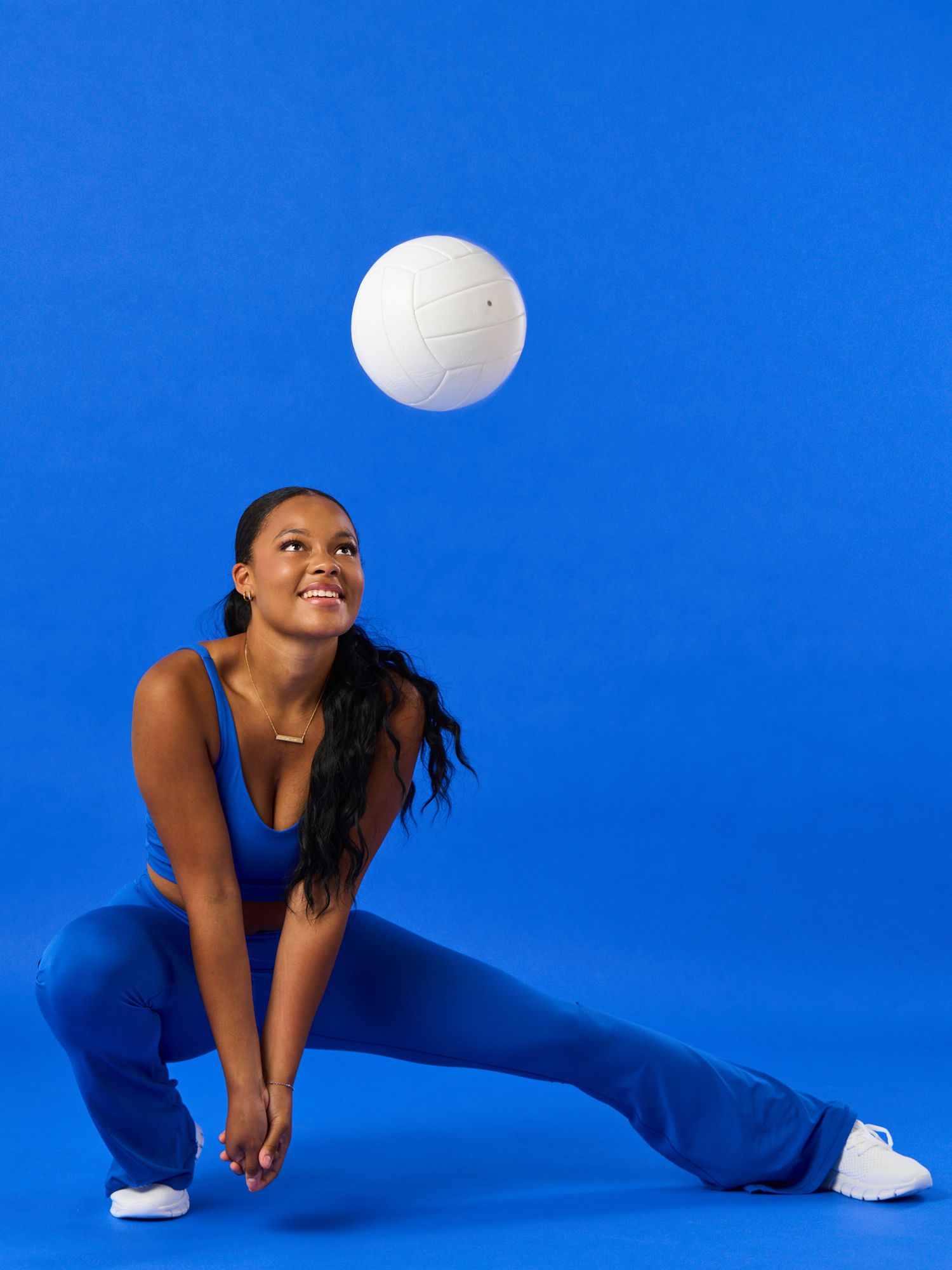 Woman in blue athletic wear lunging while bumping a white volleyball against a bright blue background.