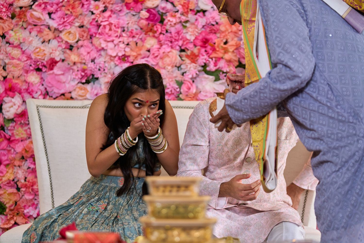 A bride in a green dress covers her mouth with bangled hands during a Hindu wedding ritual, with a pink floral wall behind her.