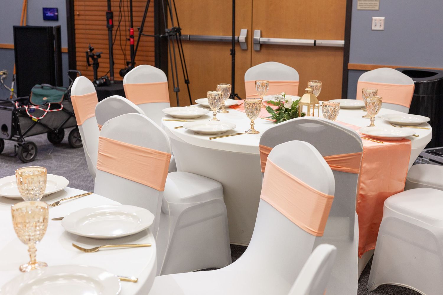 Round banquet tables set with white plates and glassware, with white chair covers tied with peach sashes in an event hall.
