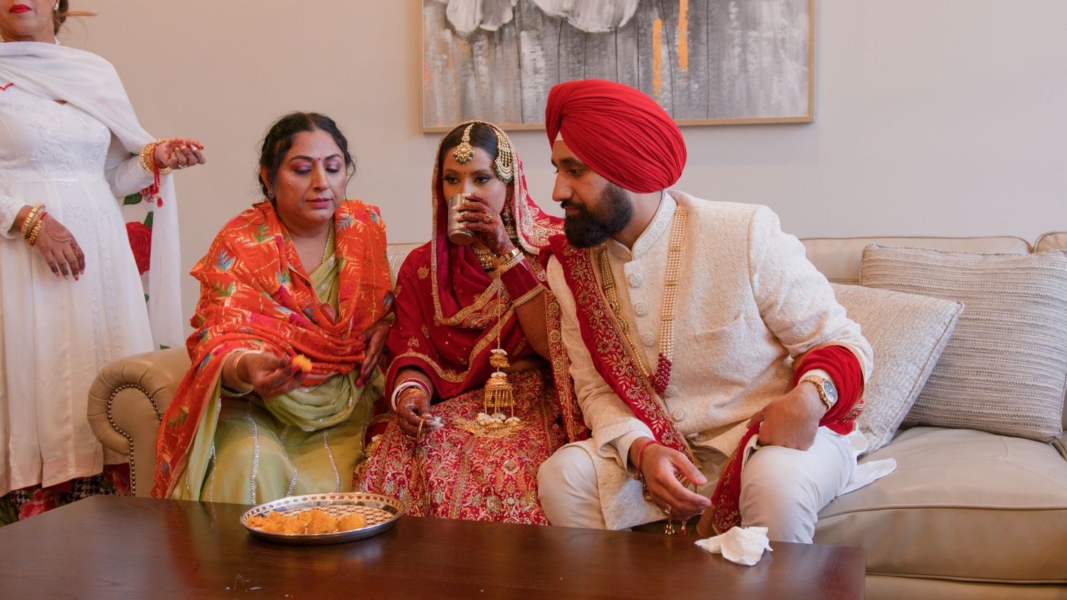 Bride in red and gold attire sits between a groom in a red turban and an older woman during a traditional Punjabi wedding ceremony.