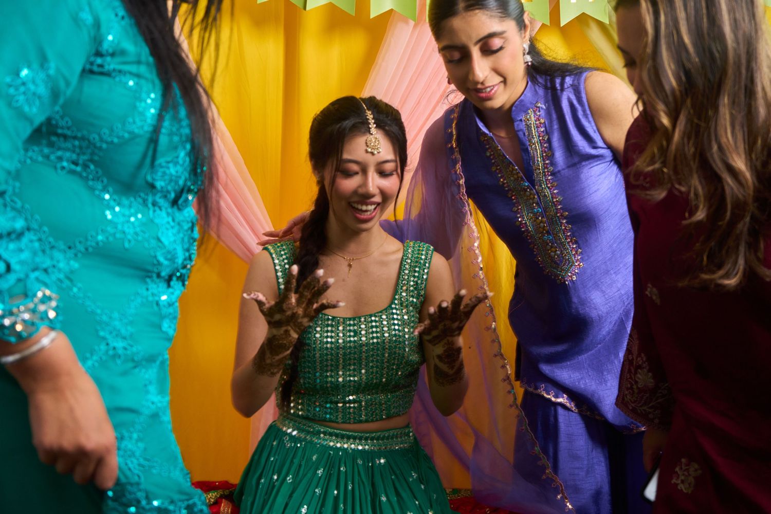 A bride in a green lehenga with mehndi hands laughs joyfully surrounded by women in colorful attire at a festive Indian celebration.