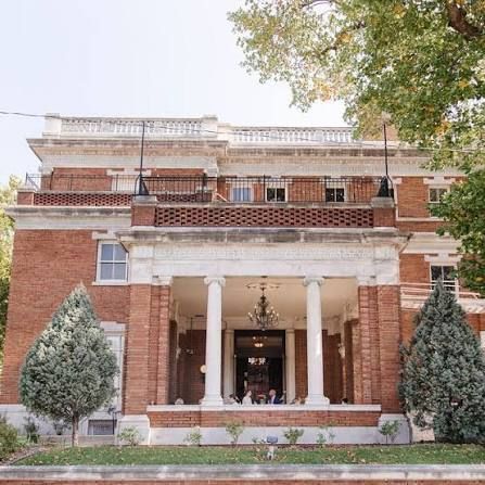 Historic red brick mansion with white columned portico, balcony, and chandelier-lit entrance framed by evergreen shrubs and mature trees.