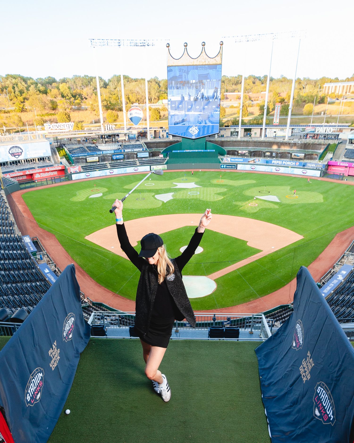 Woman celebrating with golf club raised at Kauffman Stadium baseball field during a golf event on the diamond.