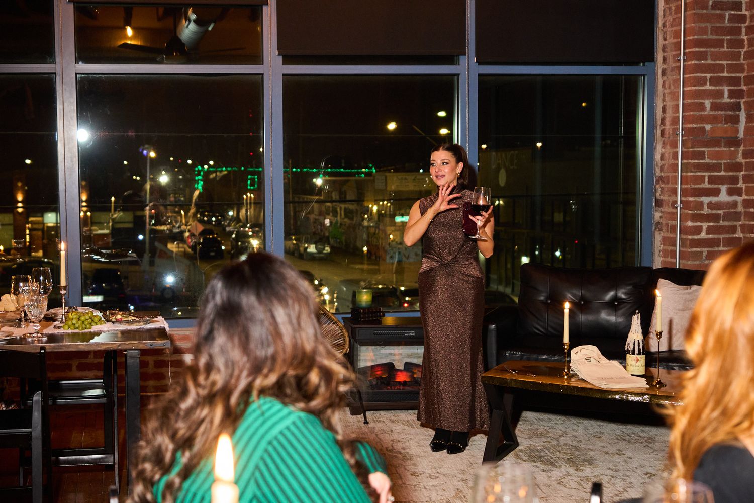 Woman in brown dress speaks at elegant rooftop dinner event with city lights visible through large windows.