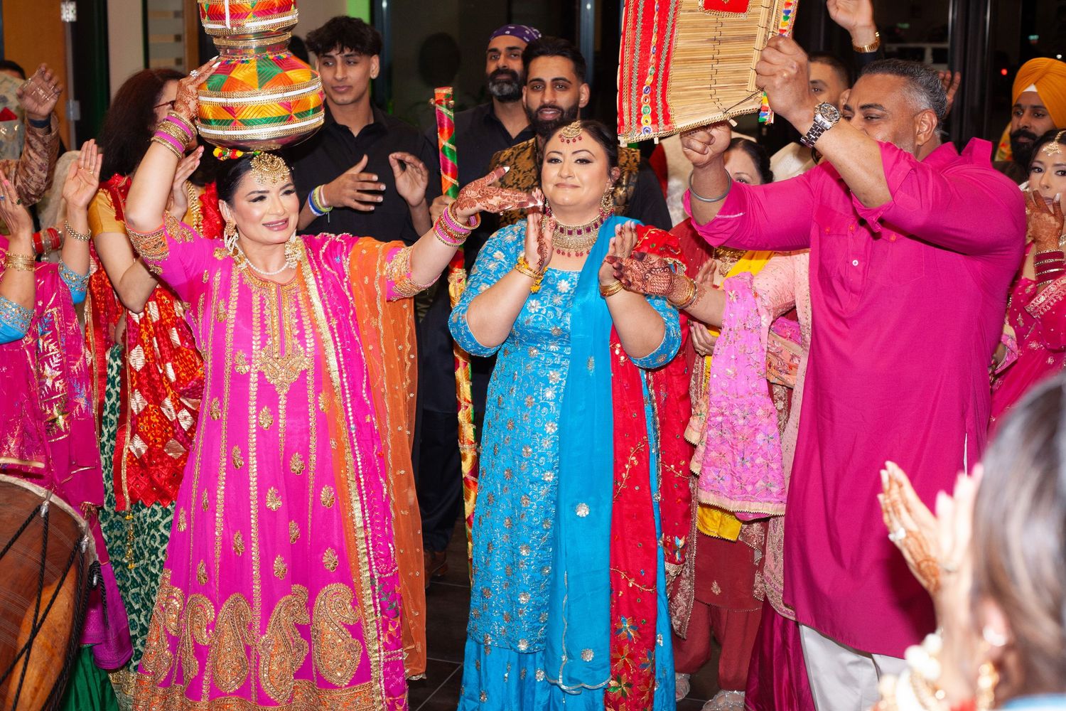Women in vibrant pink and blue traditional Indian attire clap and dance joyfully while guests celebrate at a lively wedding event.
