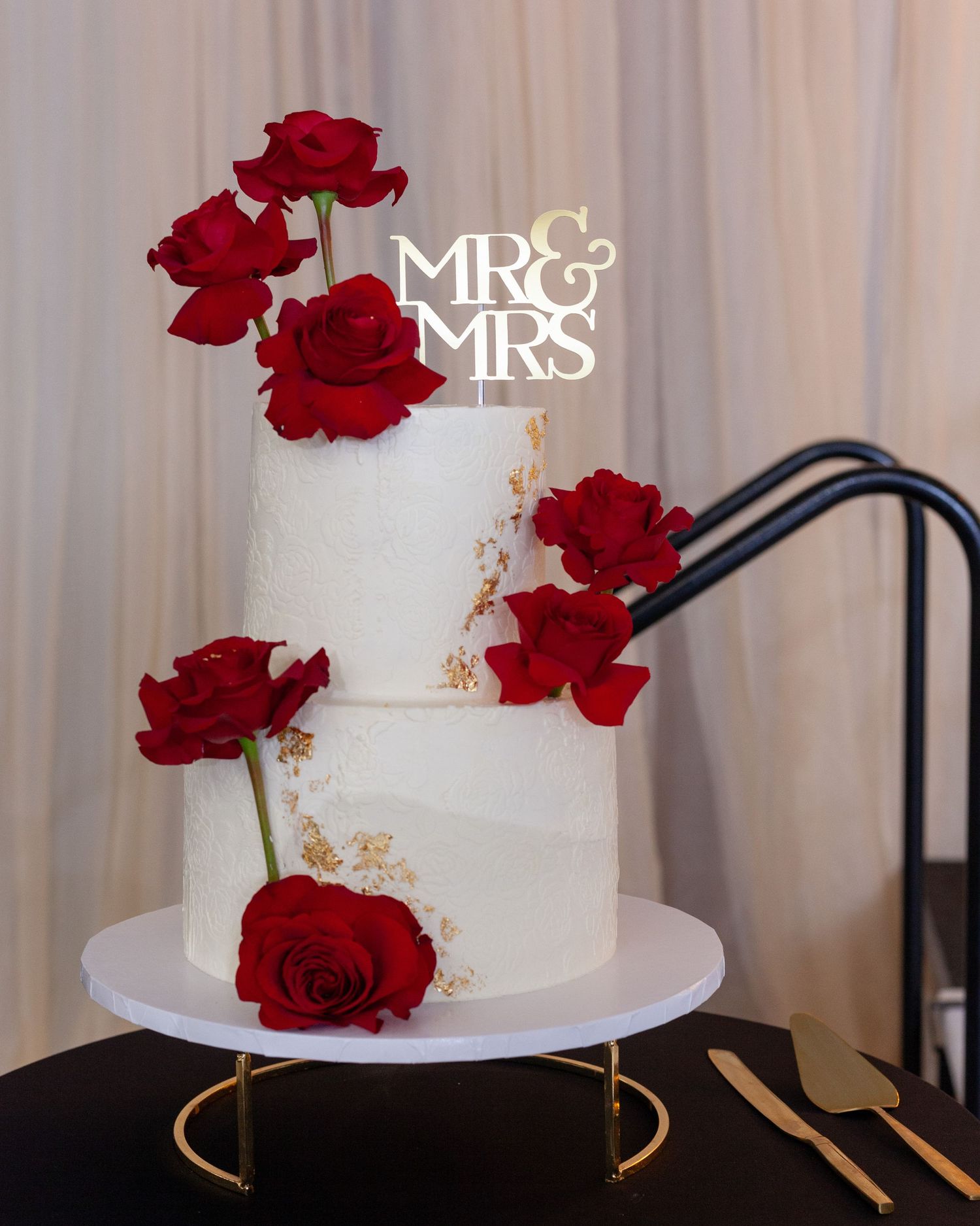 Two-tier white wedding cake with gold accents, red roses, and a Mr & Mrs topper on a white stand with gold base.