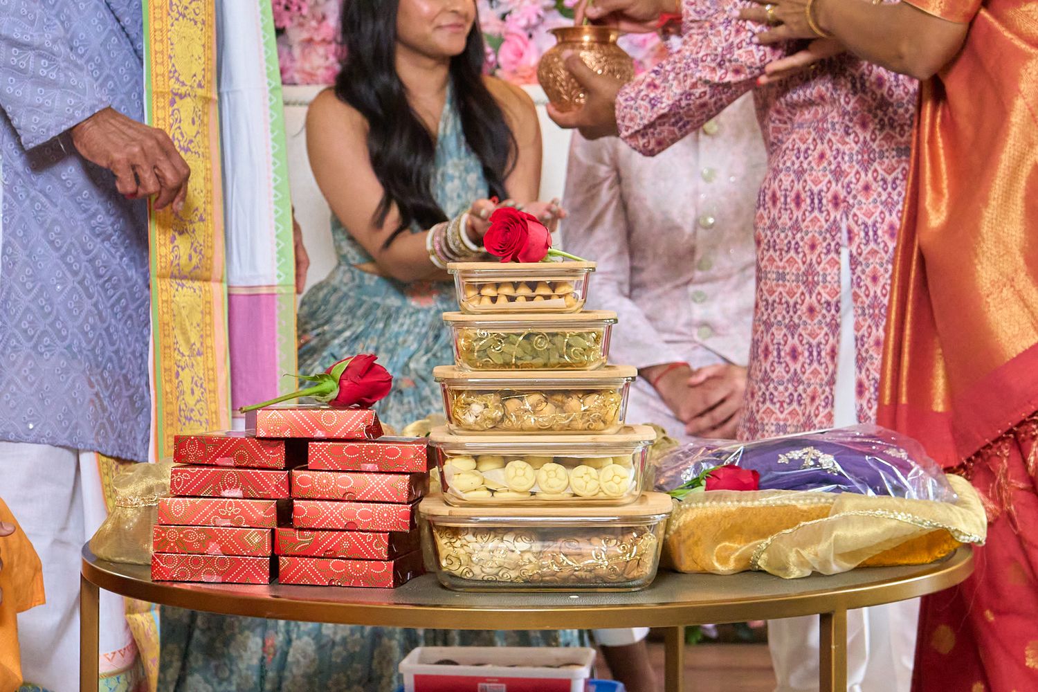 Woman in traditional attire smiles behind a table stacked with clear containers of sweets and red gift boxes at a festive celebration.