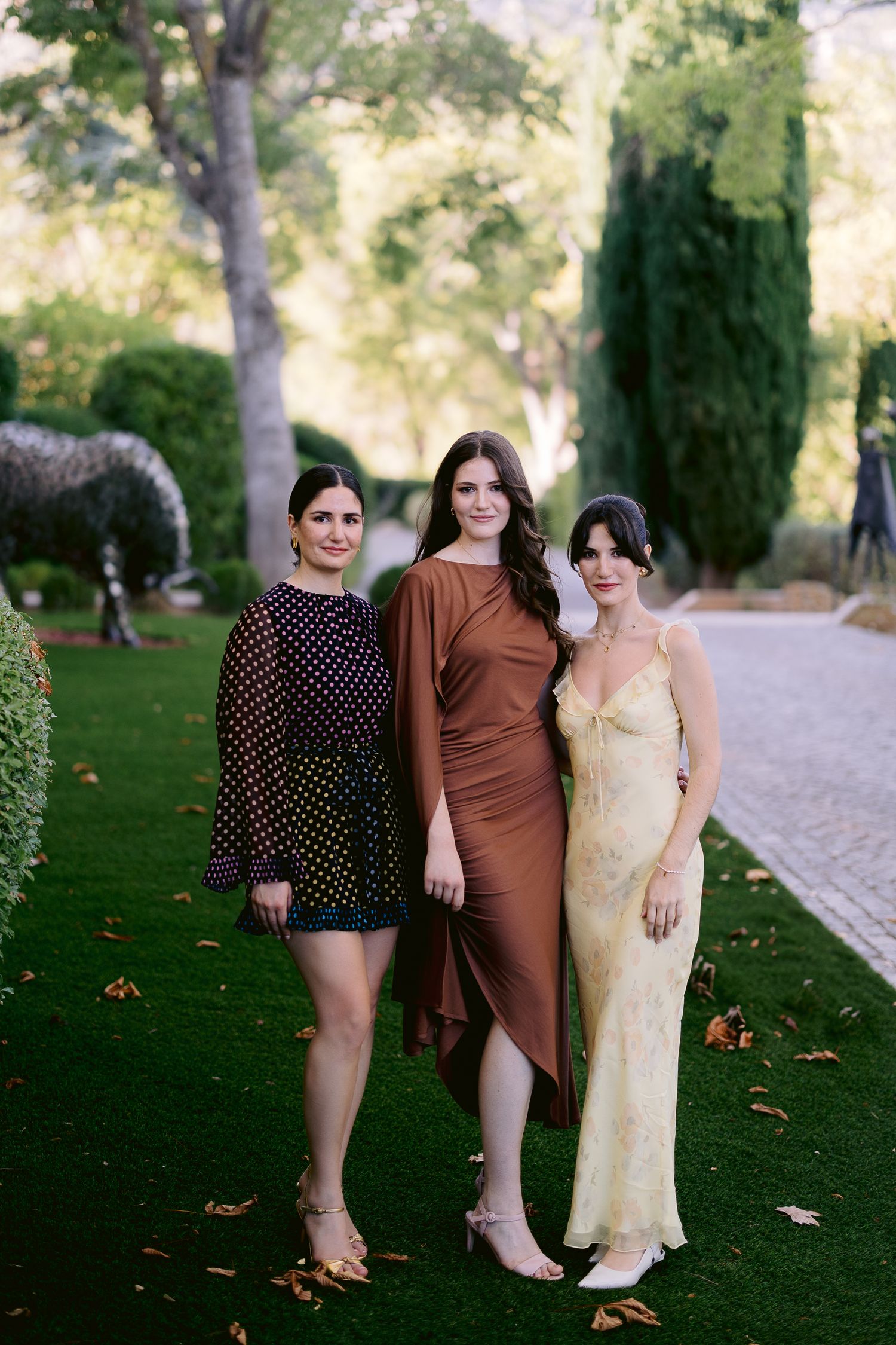 Three friends pose together on a grassy lawn in a park setting during autumn.