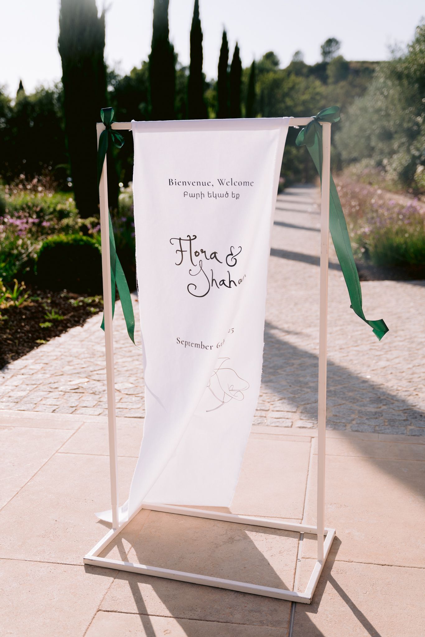 An elegant white wedding welcome banner with names and date displayed on a stand with green ribbons in an outdoor garden setting.