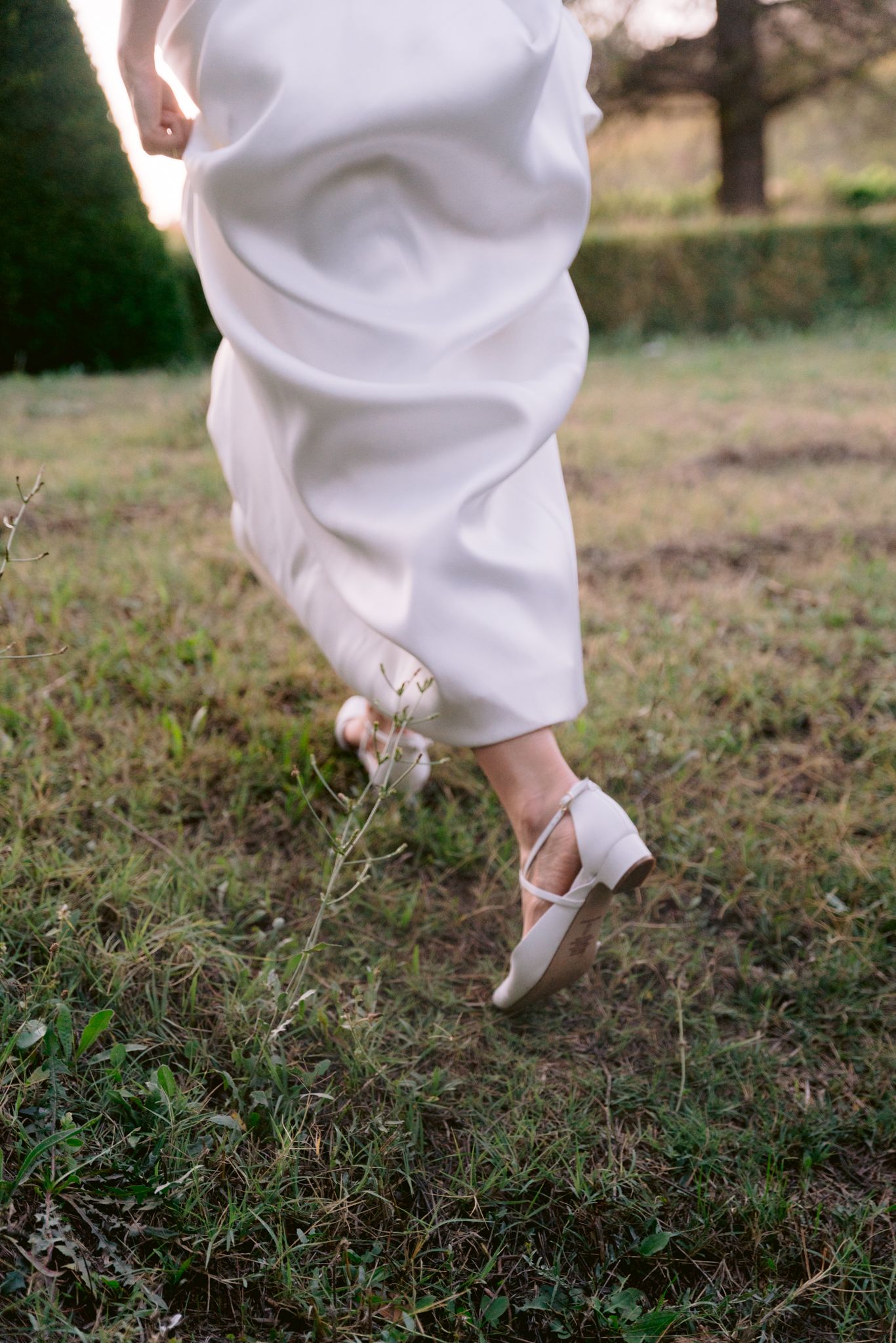 Close-up view of white wedding dress hem and shoes walking through grass in a garden setting.