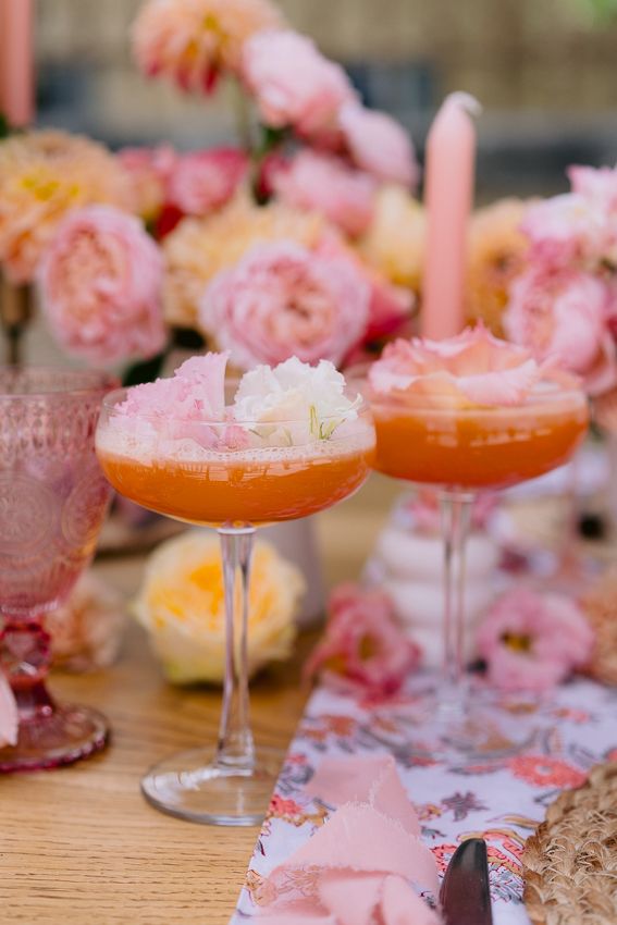 Two elegant orange cocktails garnished with flowers on a table surrounded by pink roses and candles.