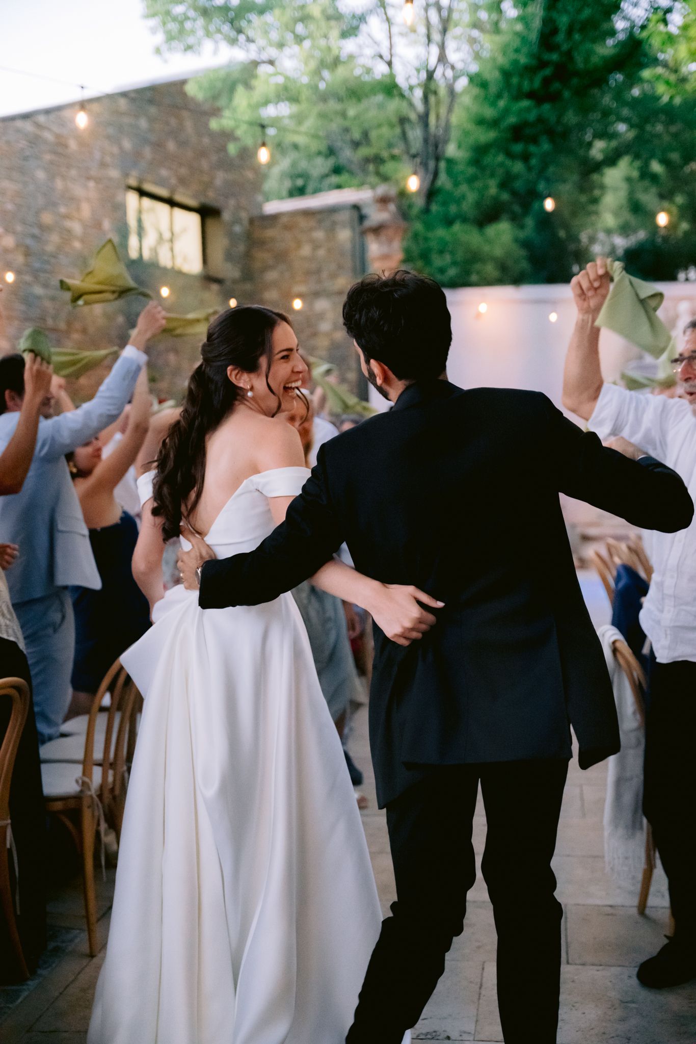 Bride in white gown smiles at groom in black suit during wedding reception exit with guests waving leaves.