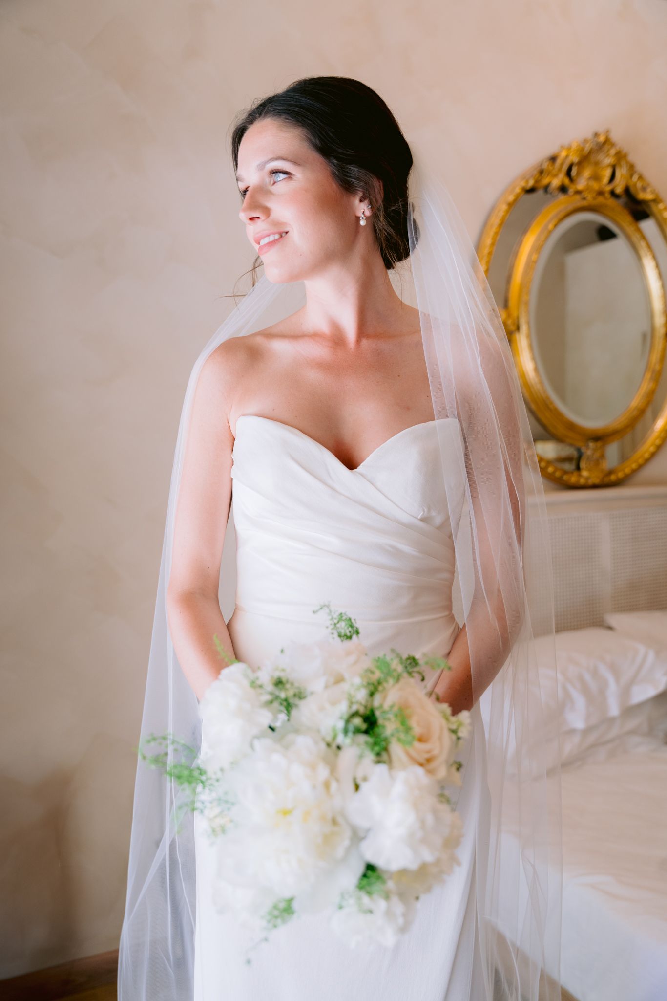 Bride in strapless white gown and veil holds white peony bouquet, smiling beside ornate gold mirror.