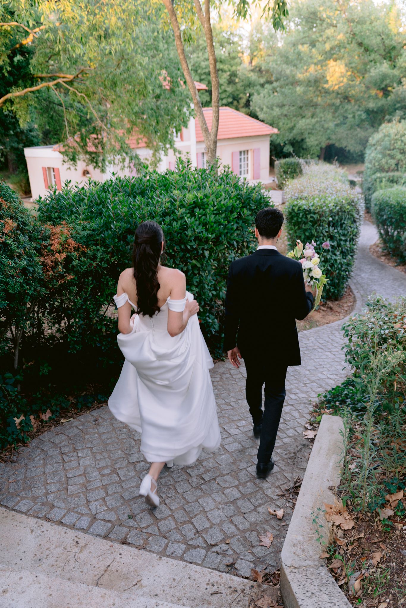 Couple walking down a winding cobblestone path surrounded by lush greenery and pink buildings at sunset.