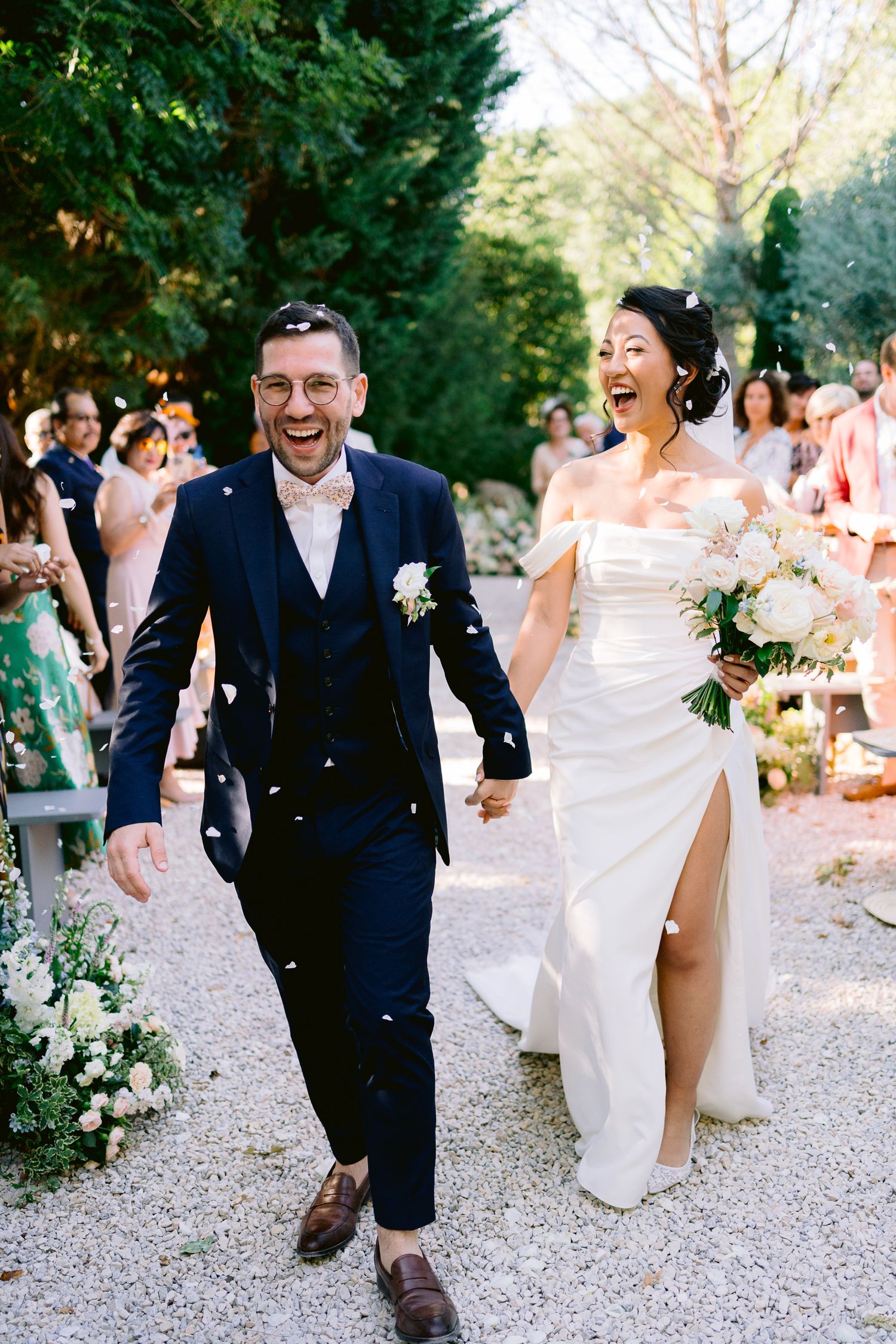Joyful newlyweds walk through confetti shower, bride in white gown holding bouquet, groom in navy suit, outdoor wedding ceremony.