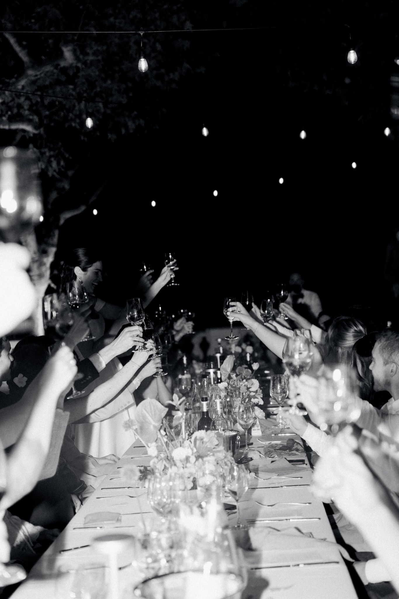 Black and white photo of a festive dinner party with guests raising glasses under twinkling lights at night.