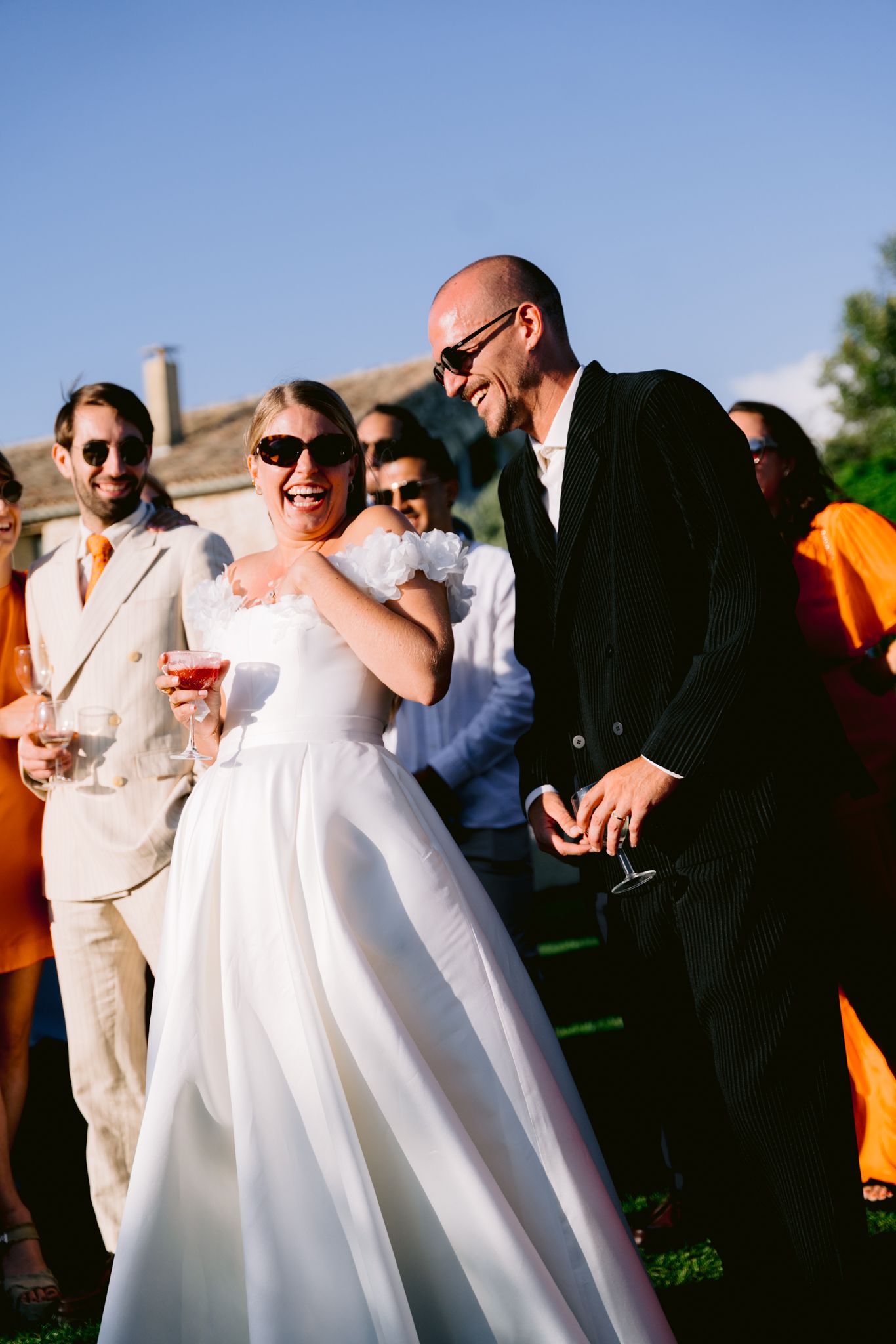Laughing bride in white gown and groom in black suit celebrate outdoors with guests at sunny wedding reception.