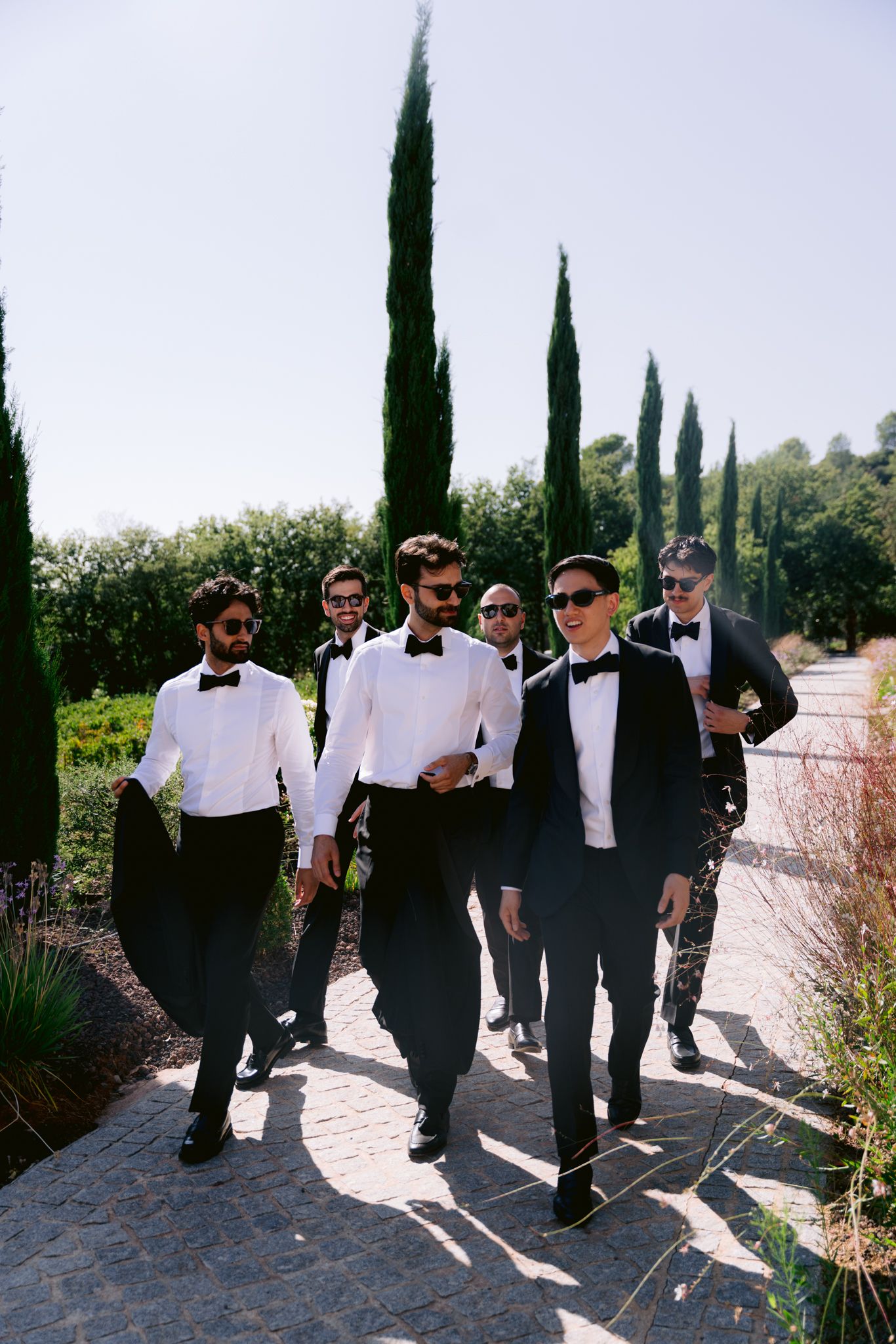 Groomsmen in black tuxedos and bow ties walk confidently along a garden path lined with tall cypress trees.