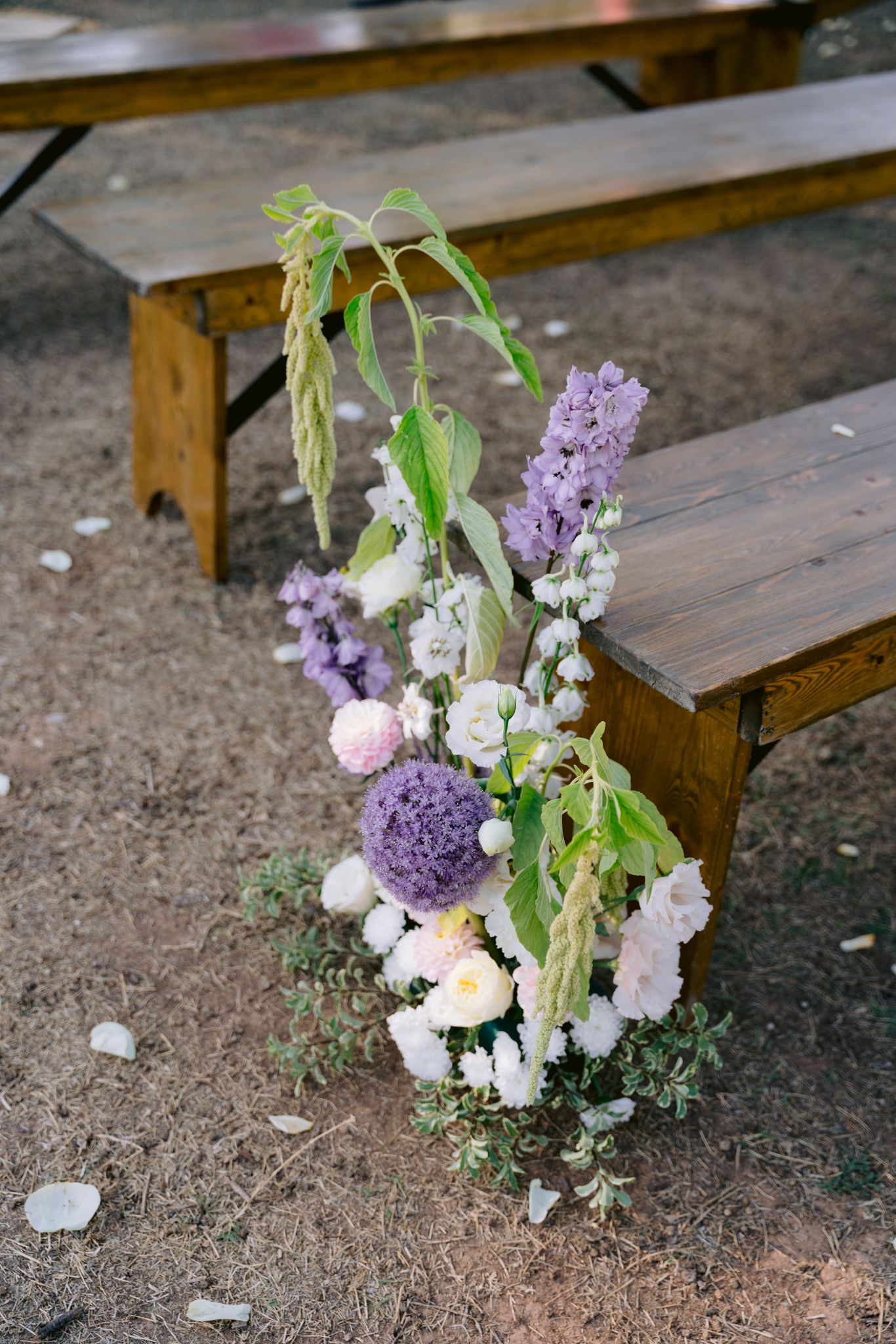 A rustic floral arrangement with purple and white blooms sits beside a wooden bench on dirt ground.