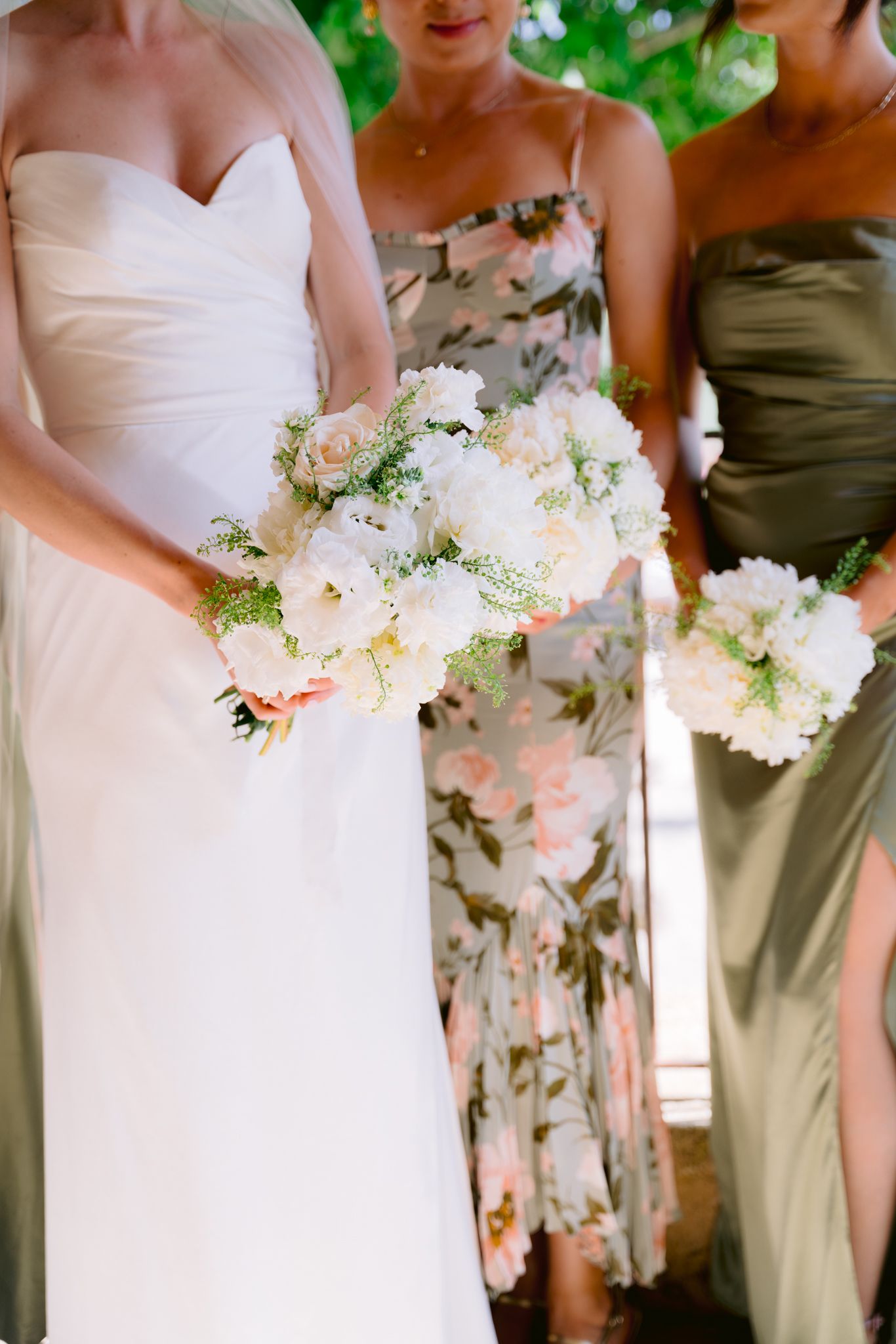 Bride and bridesmaids holding white peony bouquets, wearing white gown and floral and olive dresses.