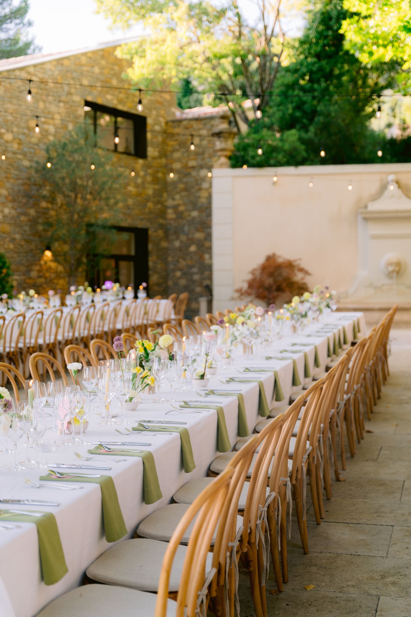 Elegant outdoor wedding reception table with long white linens, wooden chairs and stone wall backdrop in garden setting.