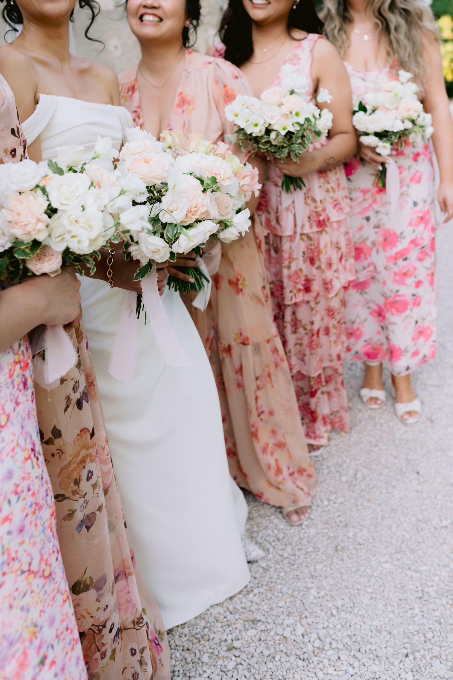 Bride and bridesmaids in floral dresses holding white and blush rose bouquets at a wedding ceremony.
