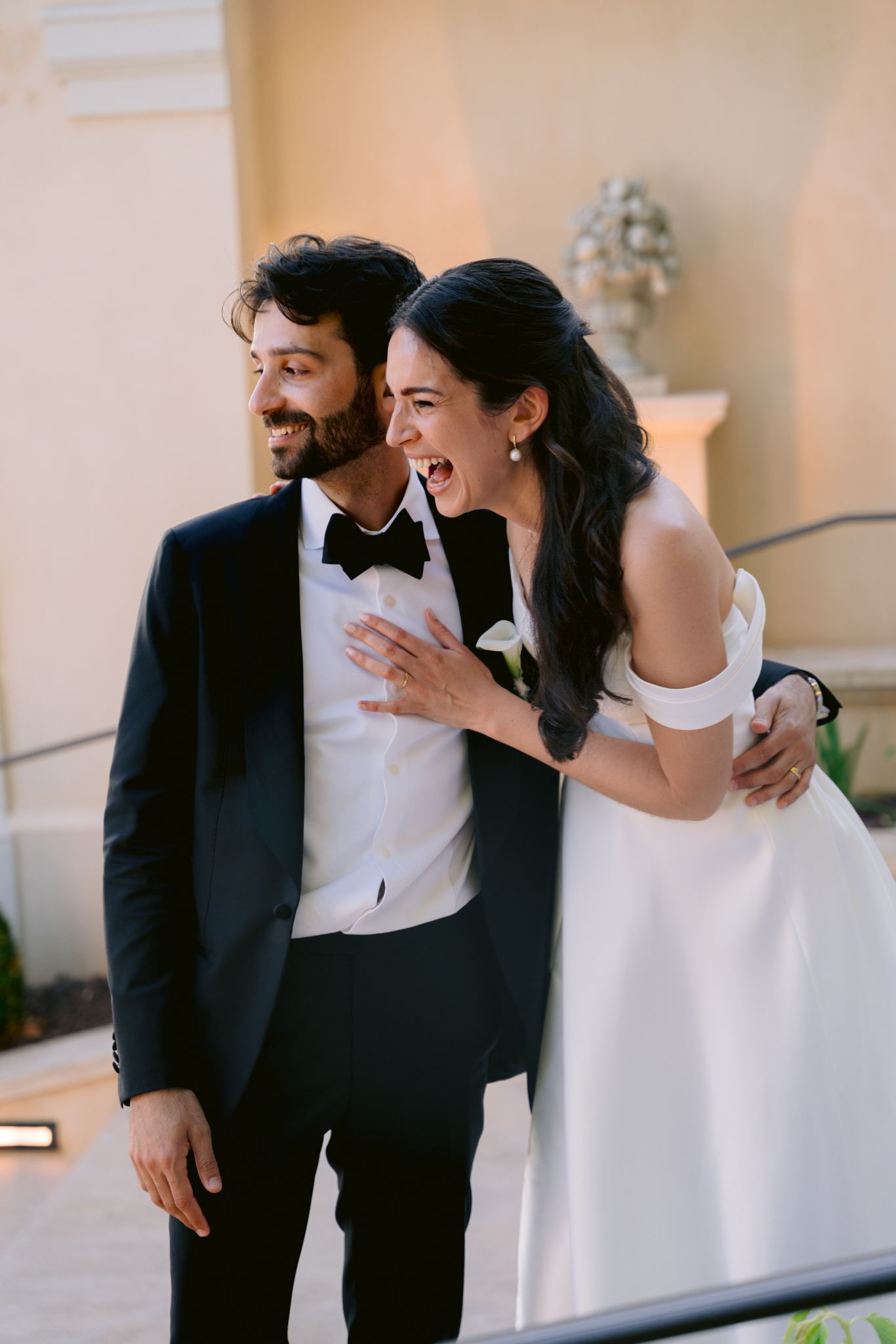 Bride and groom laughing together on steps, she wears off-shoulder white gown, he wears black tuxedo with bow tie.