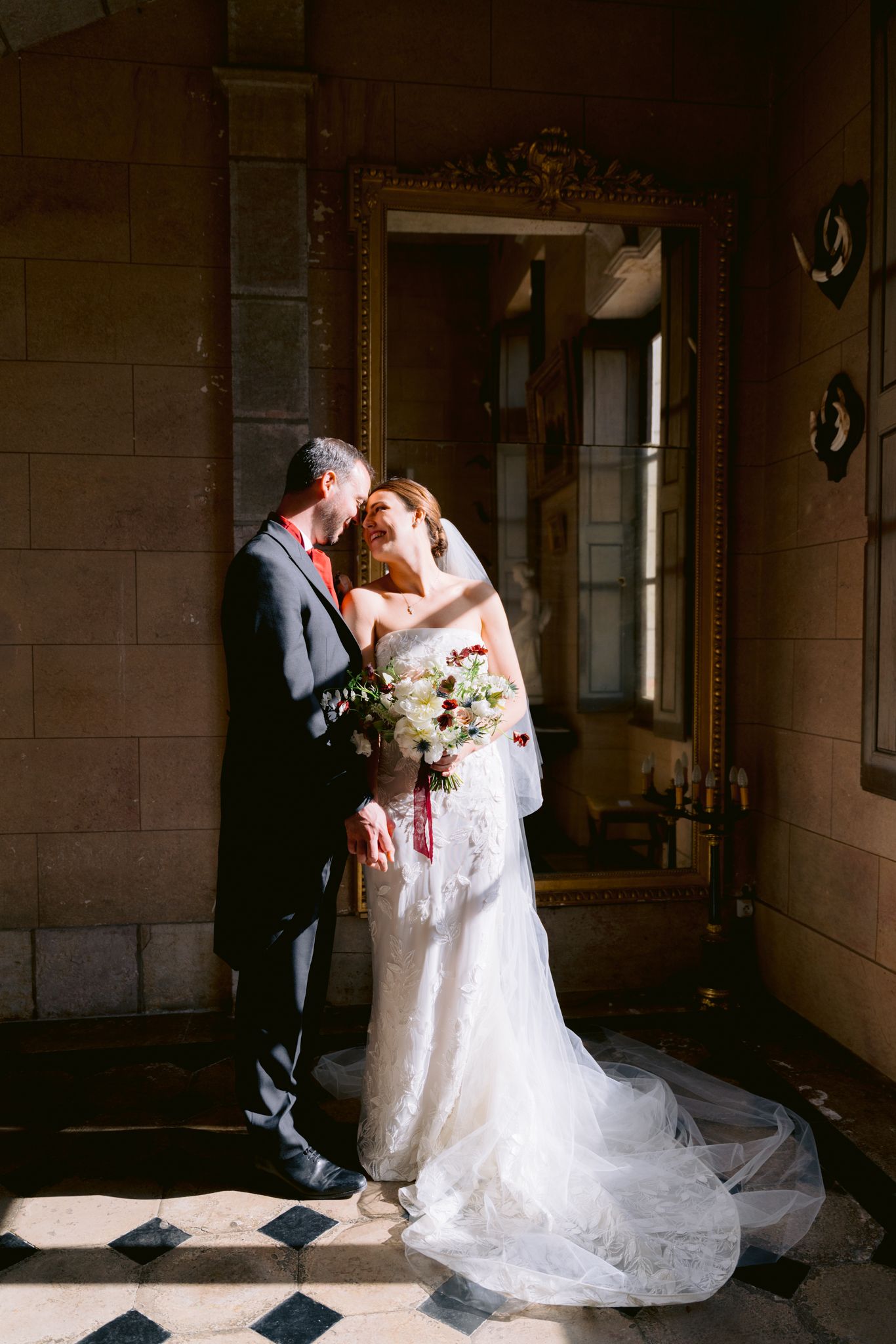 Bride and groom share a romantic kiss in a sunlit vintage hall, bride holding a bouquet in a lace gown.