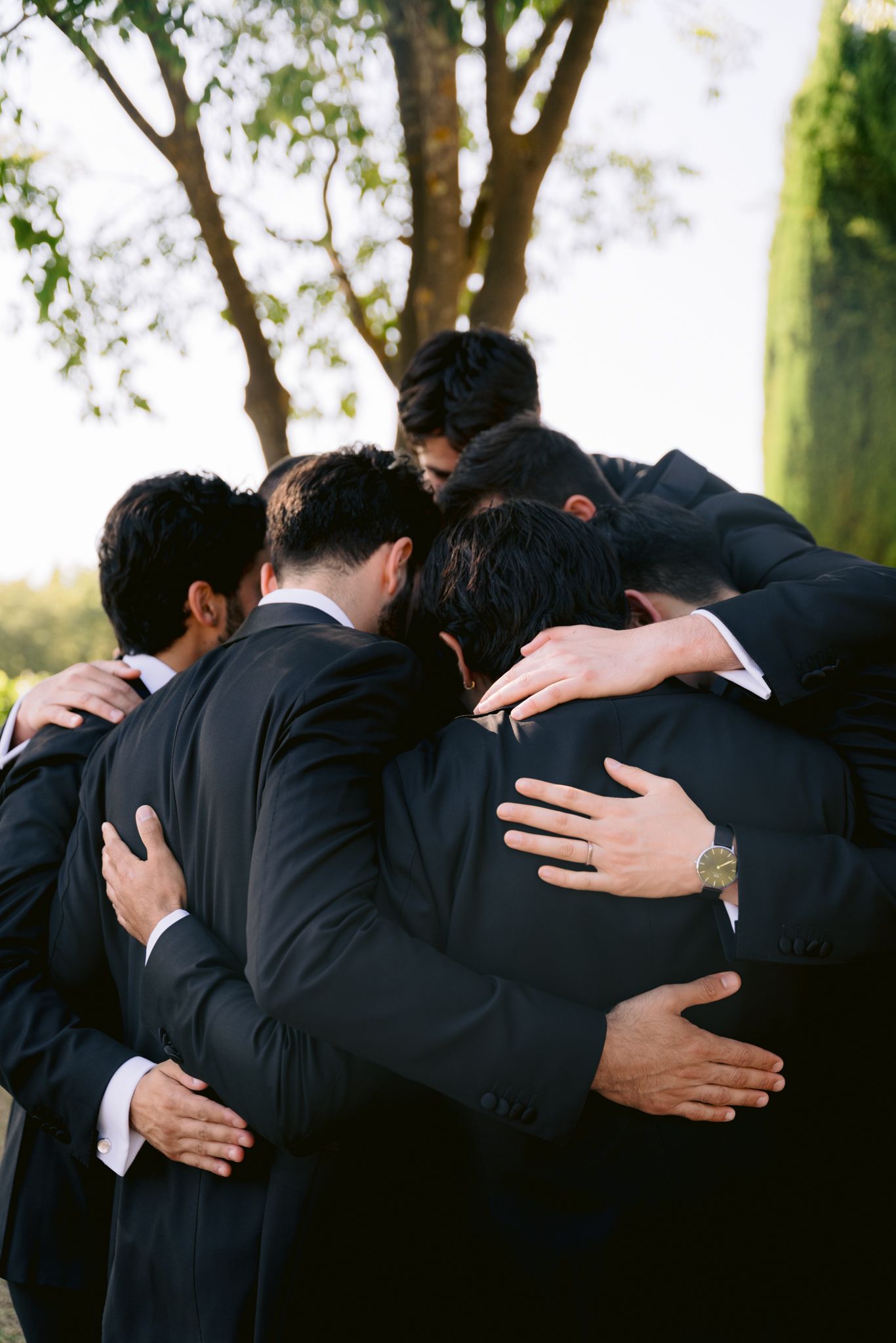 A group of men in dark suits share an emotional embrace in a circle outdoors under a tree.