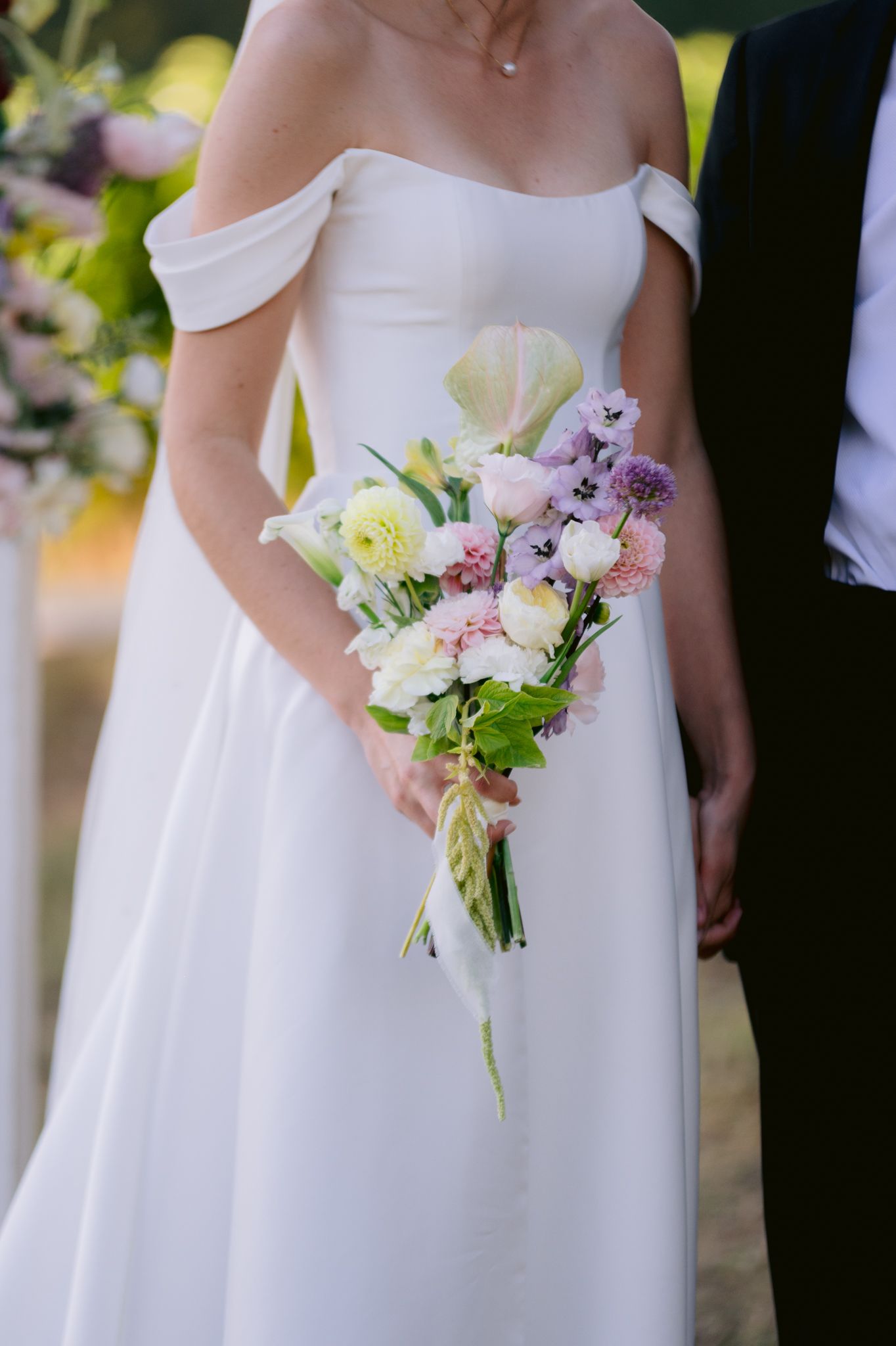 Bridesmaids in white dresses hold delicate pastel bouquets of pink and purple flowers during a wedding ceremony.
