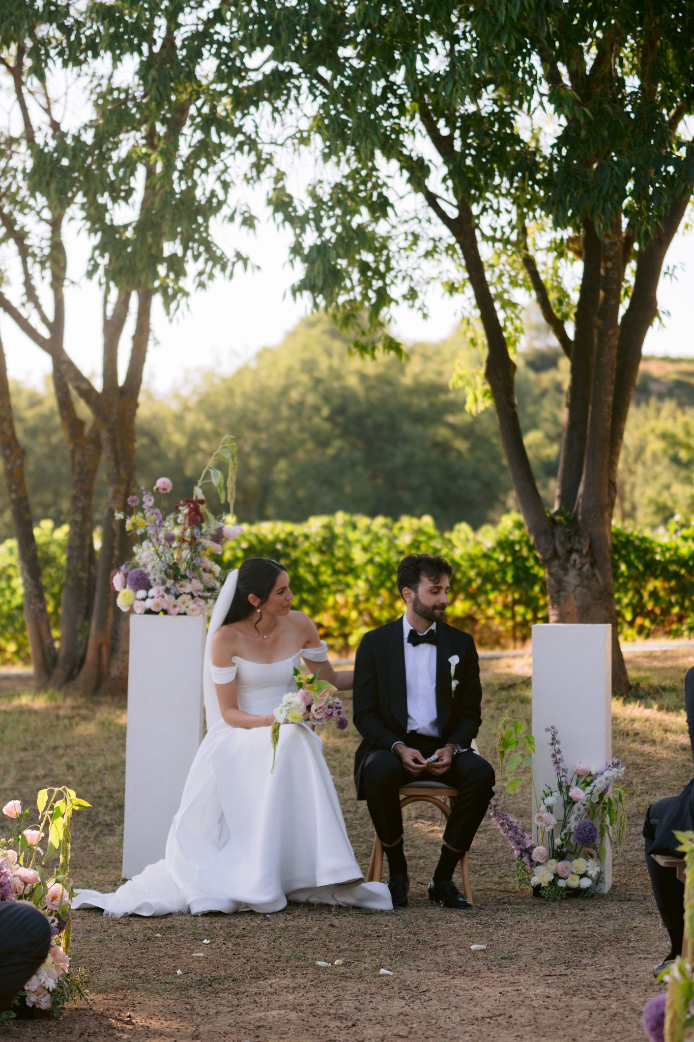 Wedding guests seated on white chairs in an outdoor garden setting with trees and natural lighting at sunset.