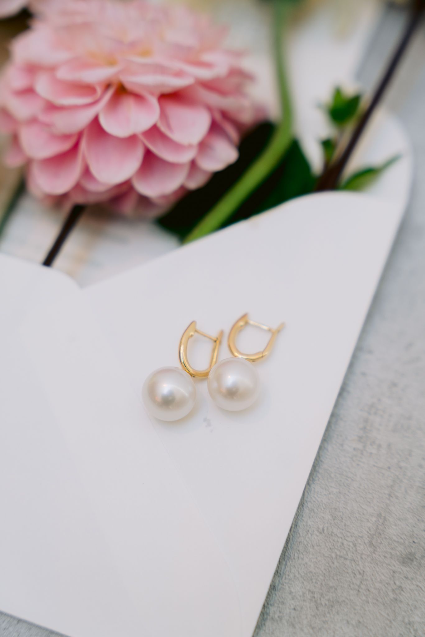 Elegant pearl drop earrings with gold hoops displayed next to a soft pink flower on white paper.