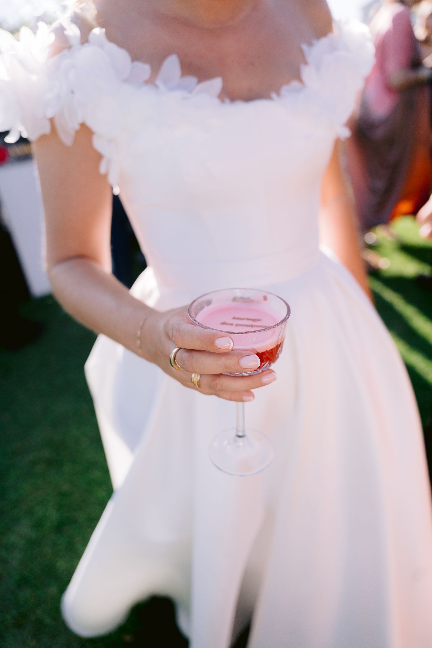 Bride in white floral gown holding a pink cocktail in a coupe glass at an outdoor wedding reception.
