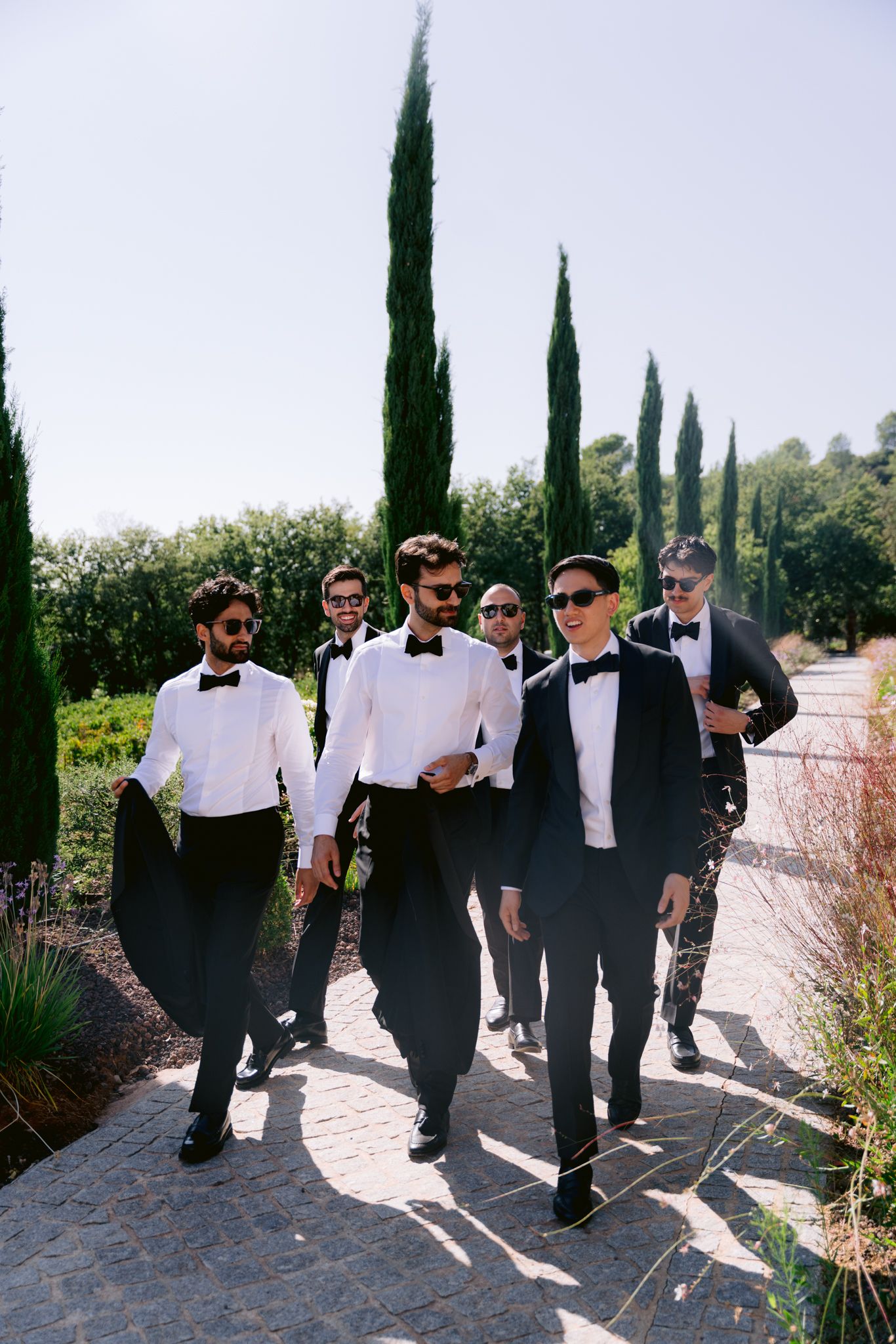 A group of groomsmen in black and white formal attire walk down a garden path lined with cypress trees.
