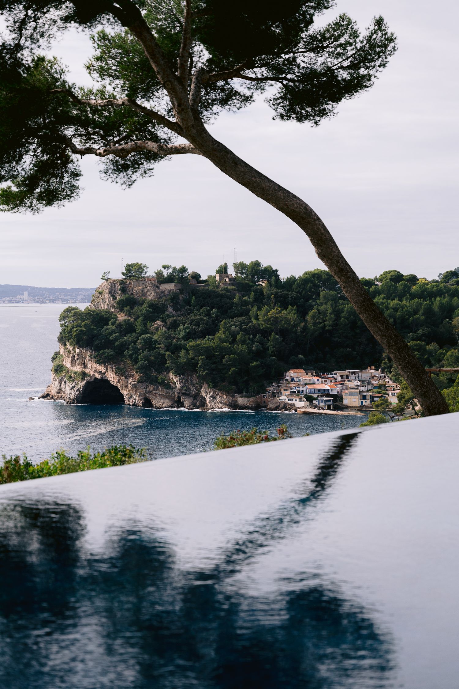 Infinity pool overlooking a rocky Mediterranean coastal village with lush green cliffs and calm sea under overcast skies.