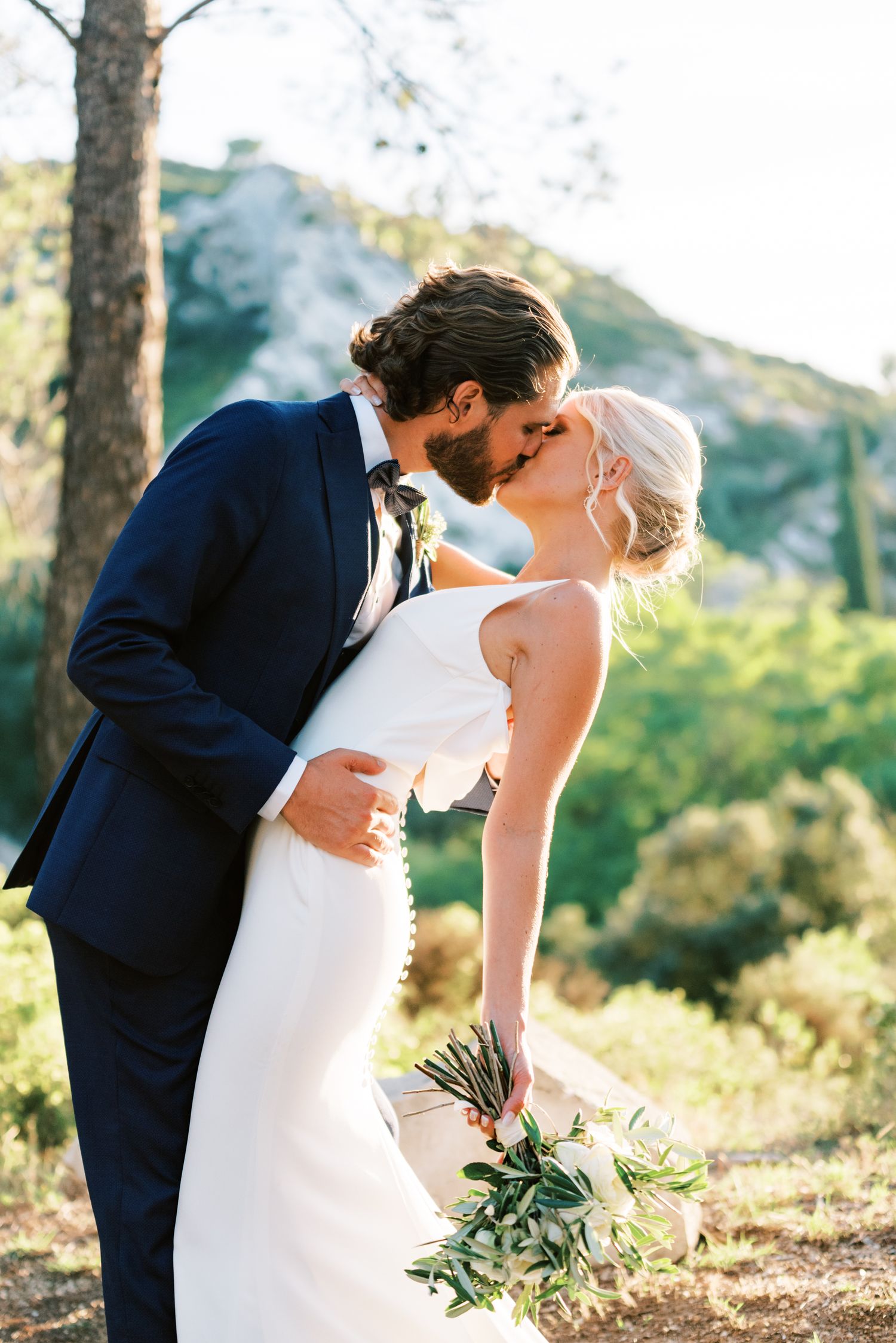 Bride and groom share a romantic kiss outdoors, she holds a greenery bouquet, he wears a navy suit, mountains in background.