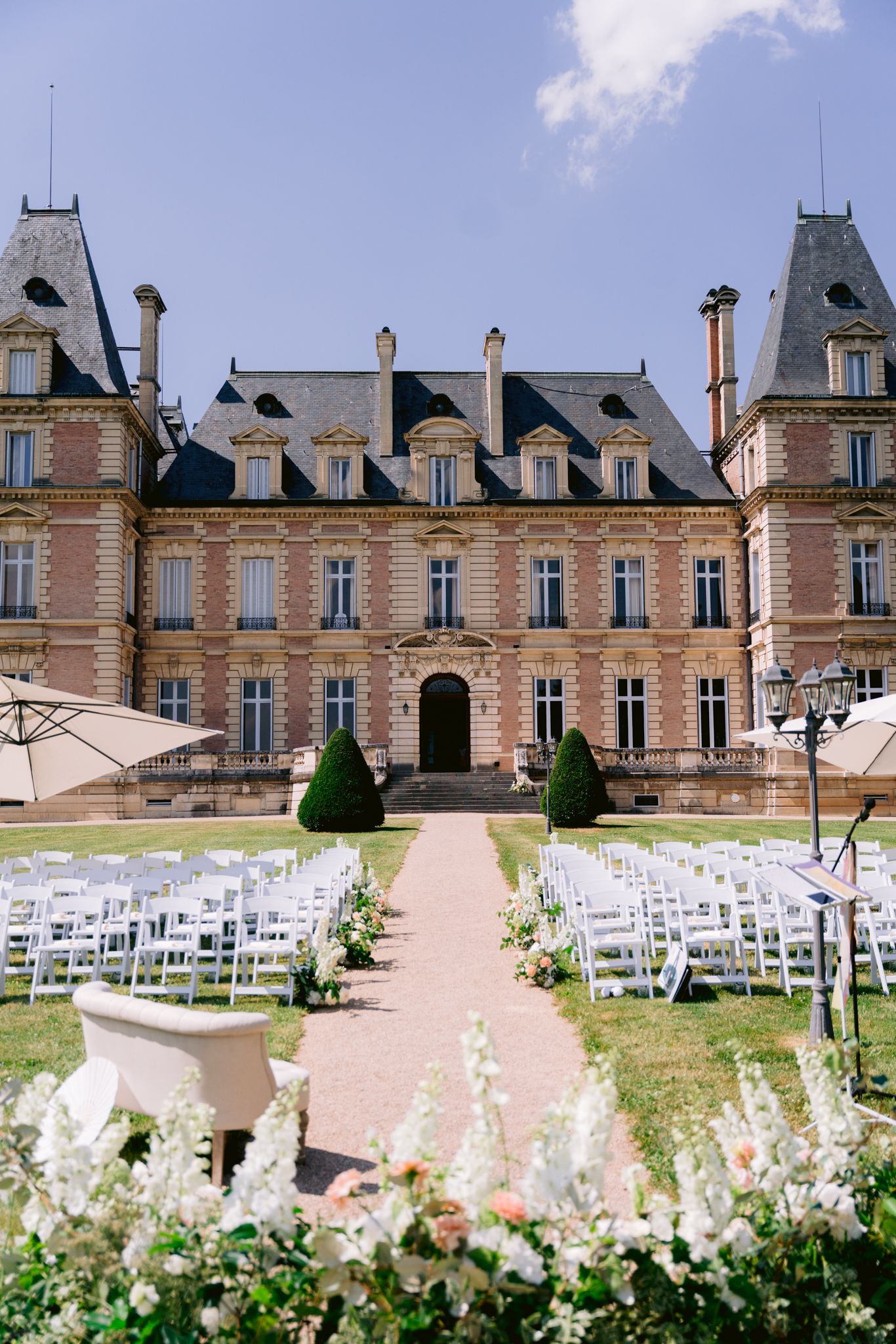 Elegant French chateau wedding ceremony setup with white chairs, floral arrangements, and manicured gardens under blue sky.
