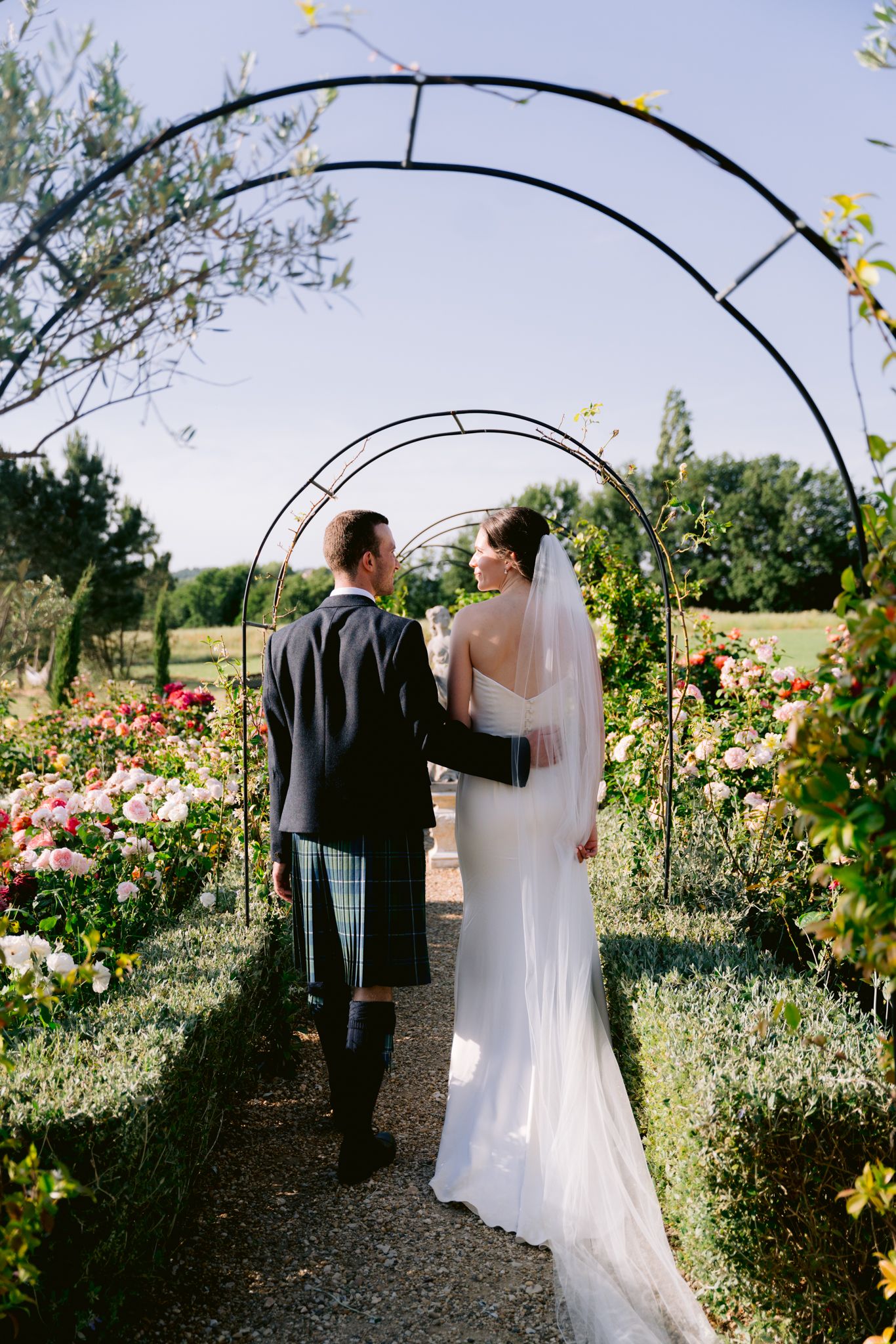 Bride and groom walk through rose garden arch, bride in white gown with veil, groom in Scottish kilt.