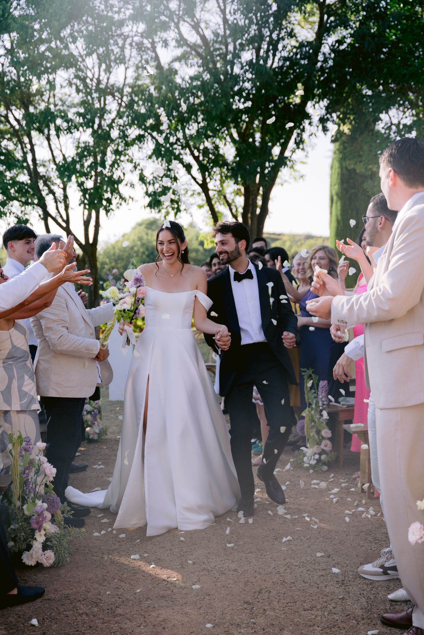 Bride and groom walk joyfully through petal toss after outdoor wedding ceremony, surrounded by cheering guests under trees.
