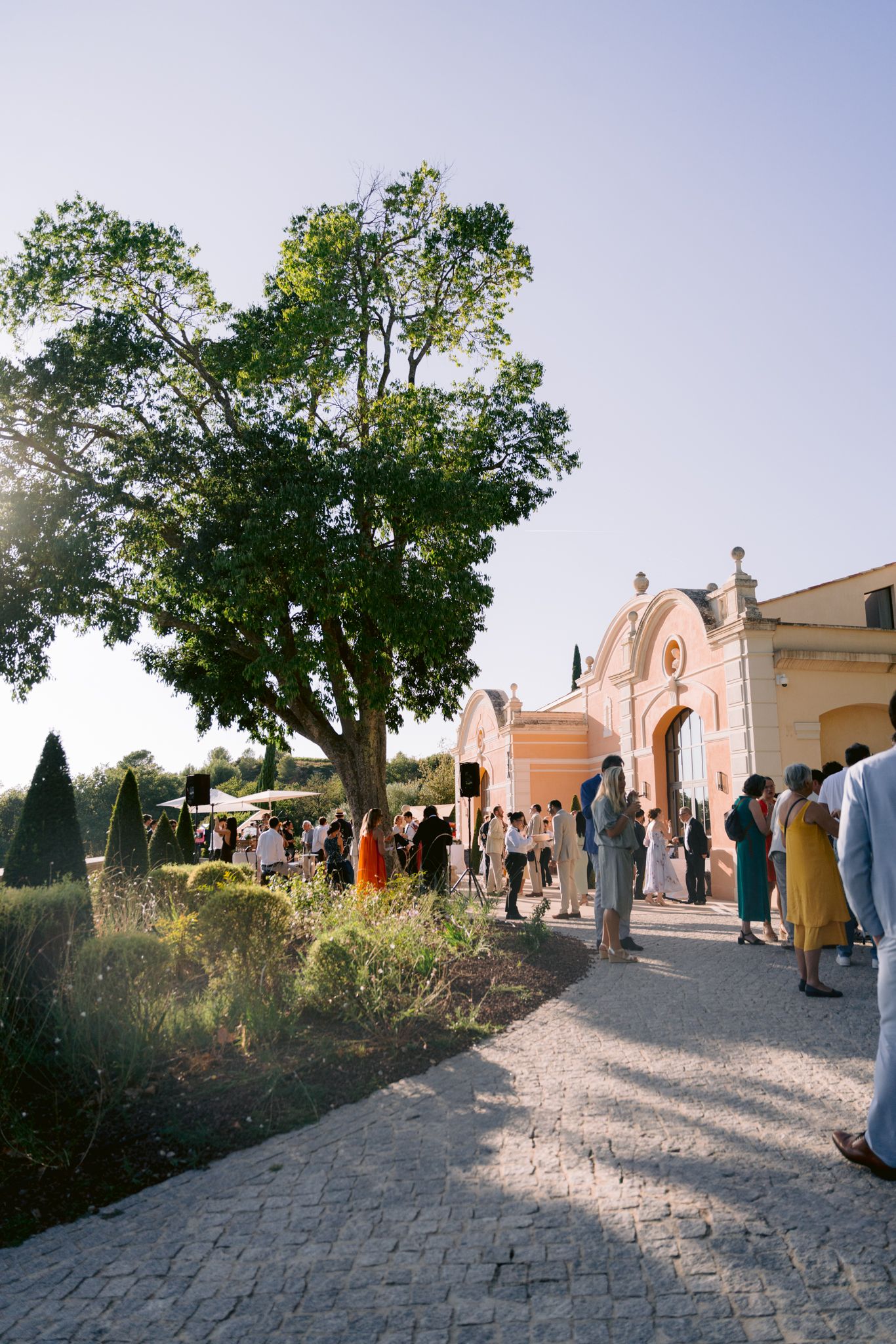 People gather outside a pink-colored church building with arched doorways on a sunny afternoon in a rustic setting.