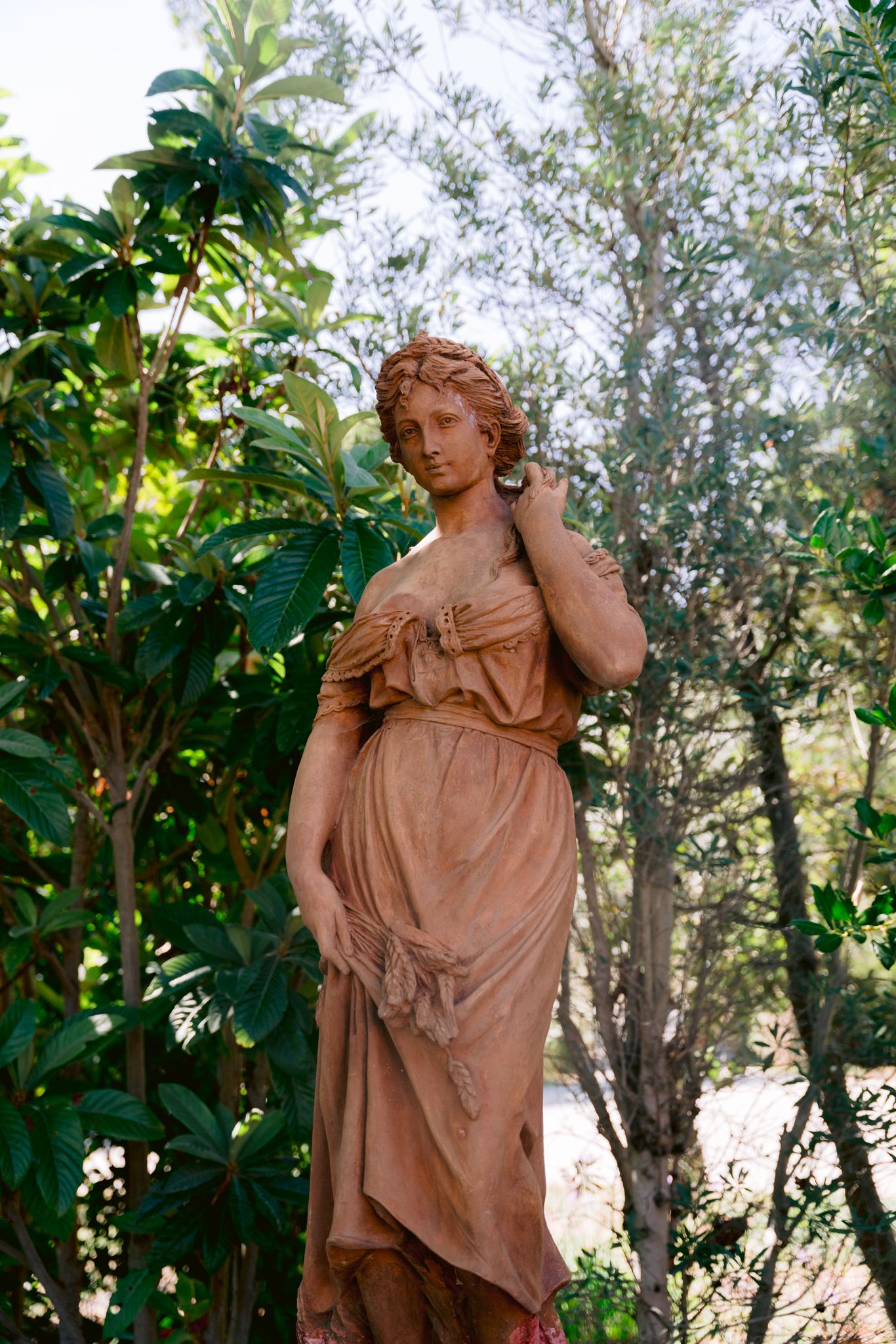 Classical terracotta garden statue of a draped female figure surrounded by lush greenery and trees.