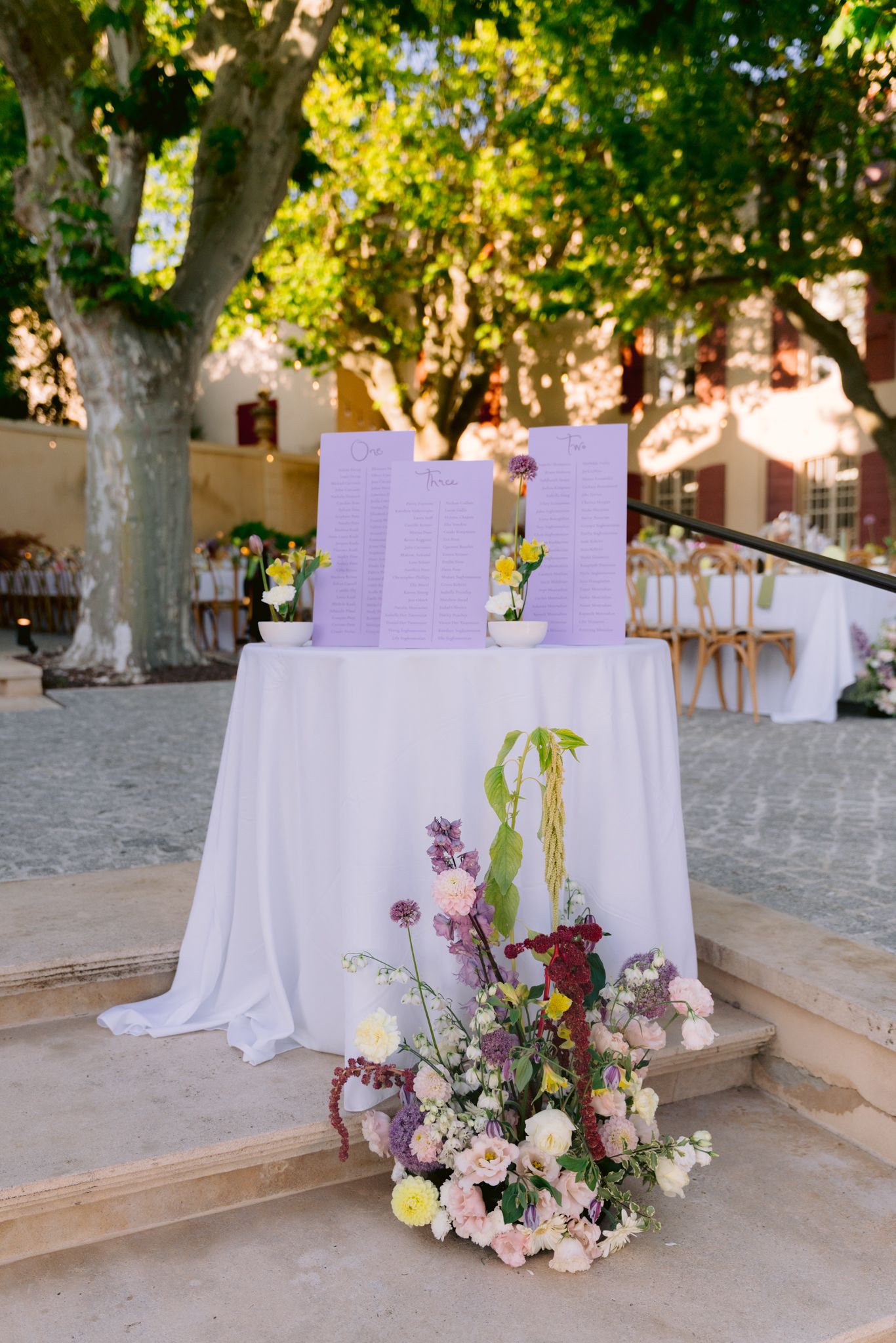 A series of wedding ceremony tables with purple stationery and floral arrangements set against a rustic outdoor backdrop.