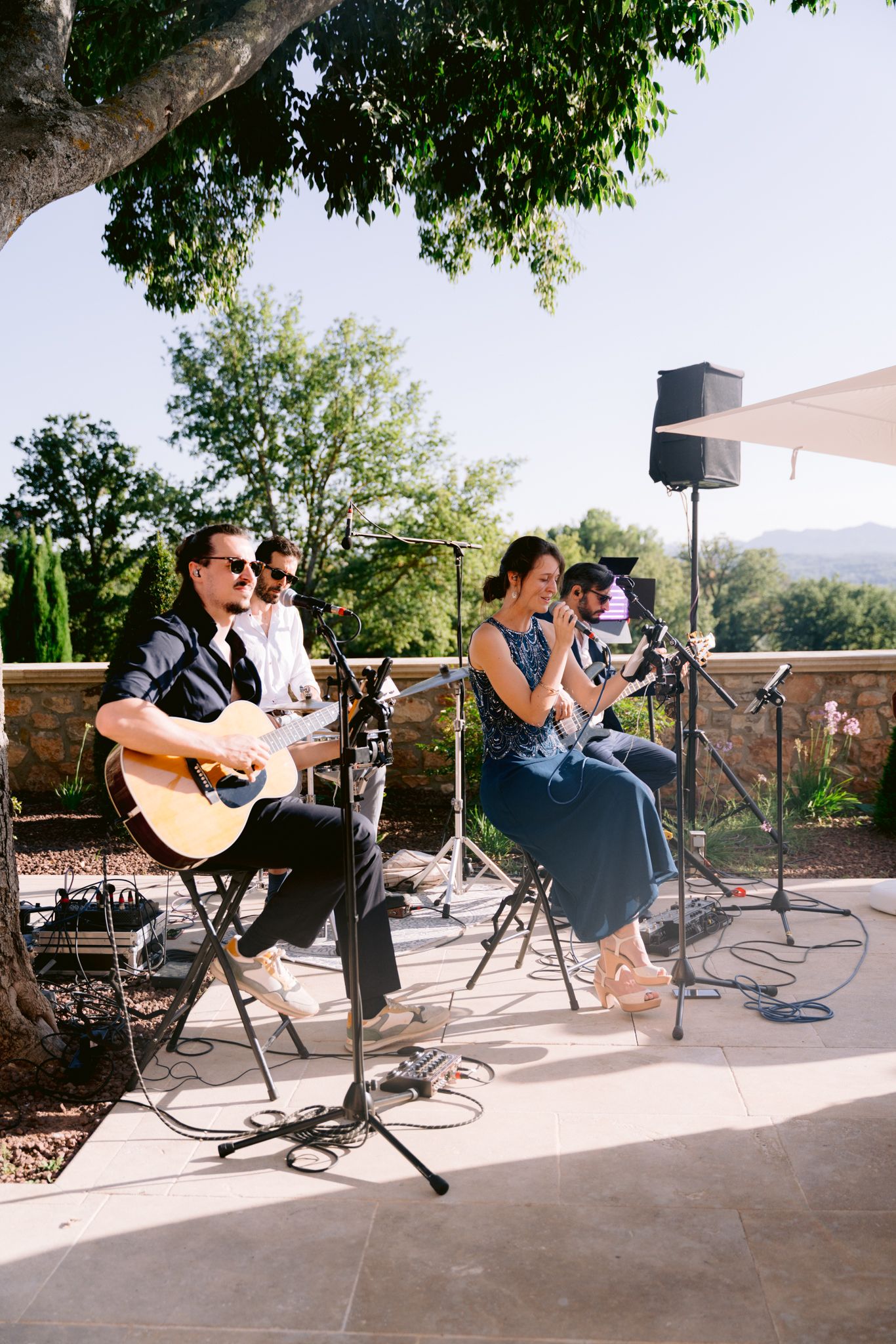 Musicians perform on an outdoor patio stage with guitars and microphones under tree cover.