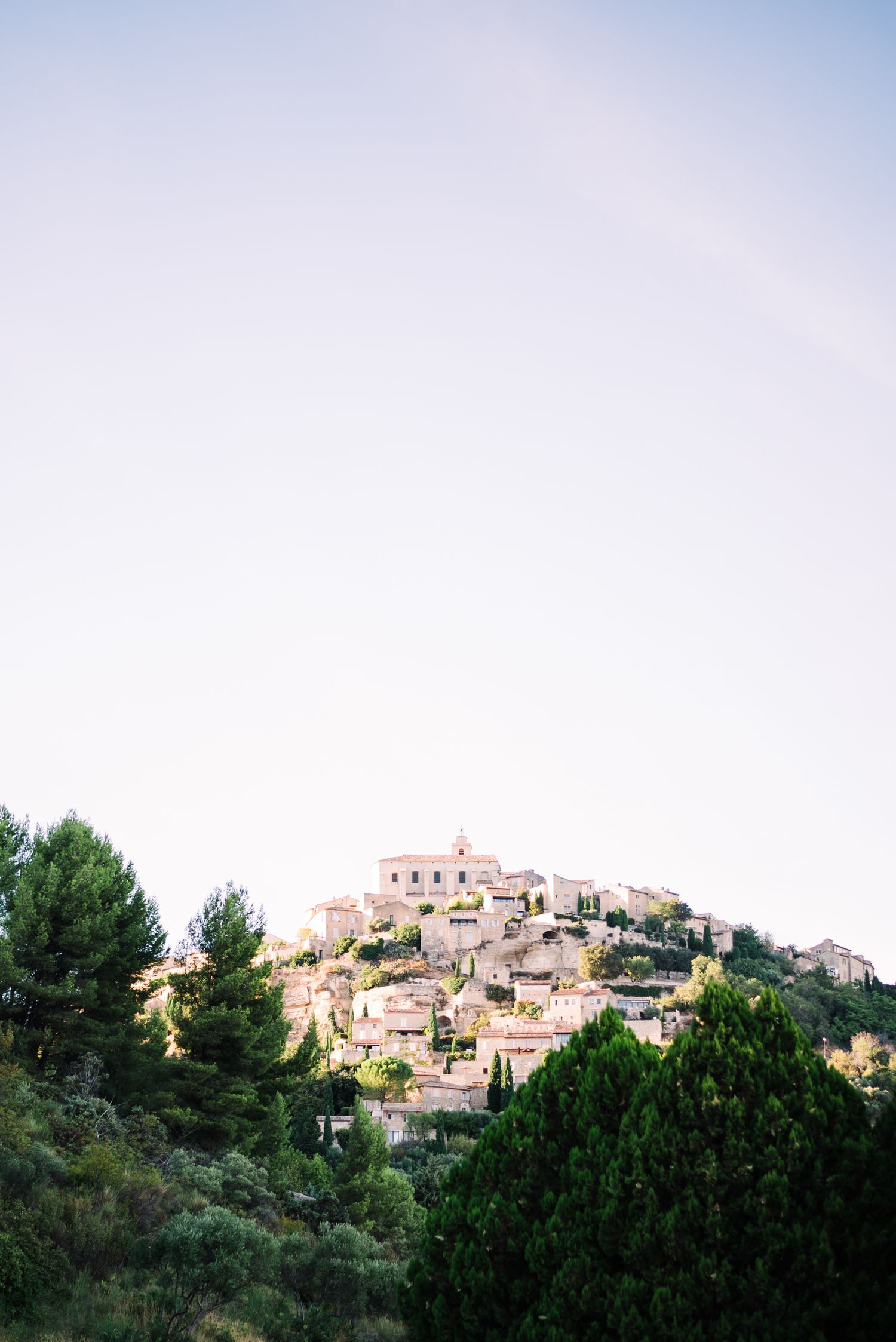 A hilltop village with stone buildings surrounded by lush green trees under a soft pastel sky.